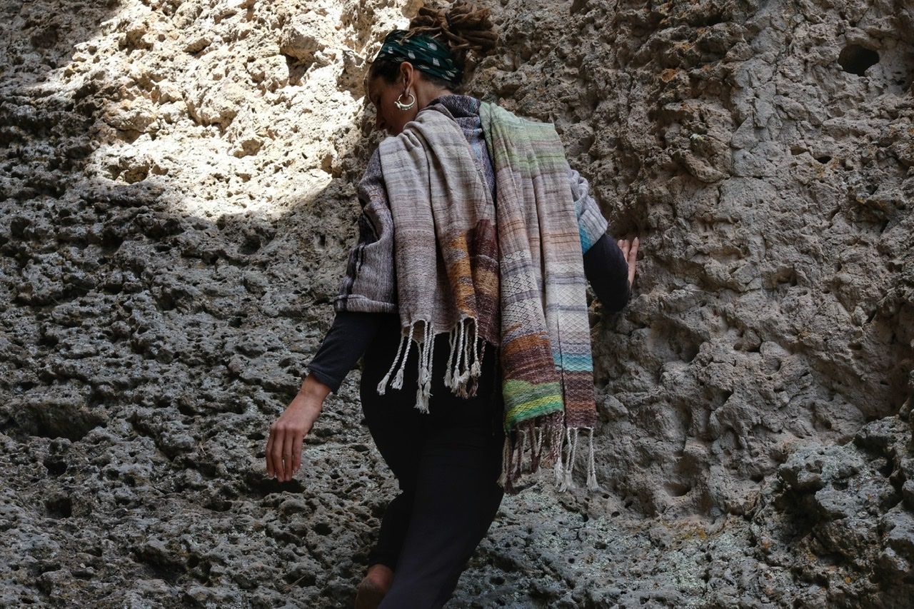 A woman wears a handwoven, diamond pattern shawl in blues, grey, black, brown, pink and green while standing in a stone canyon