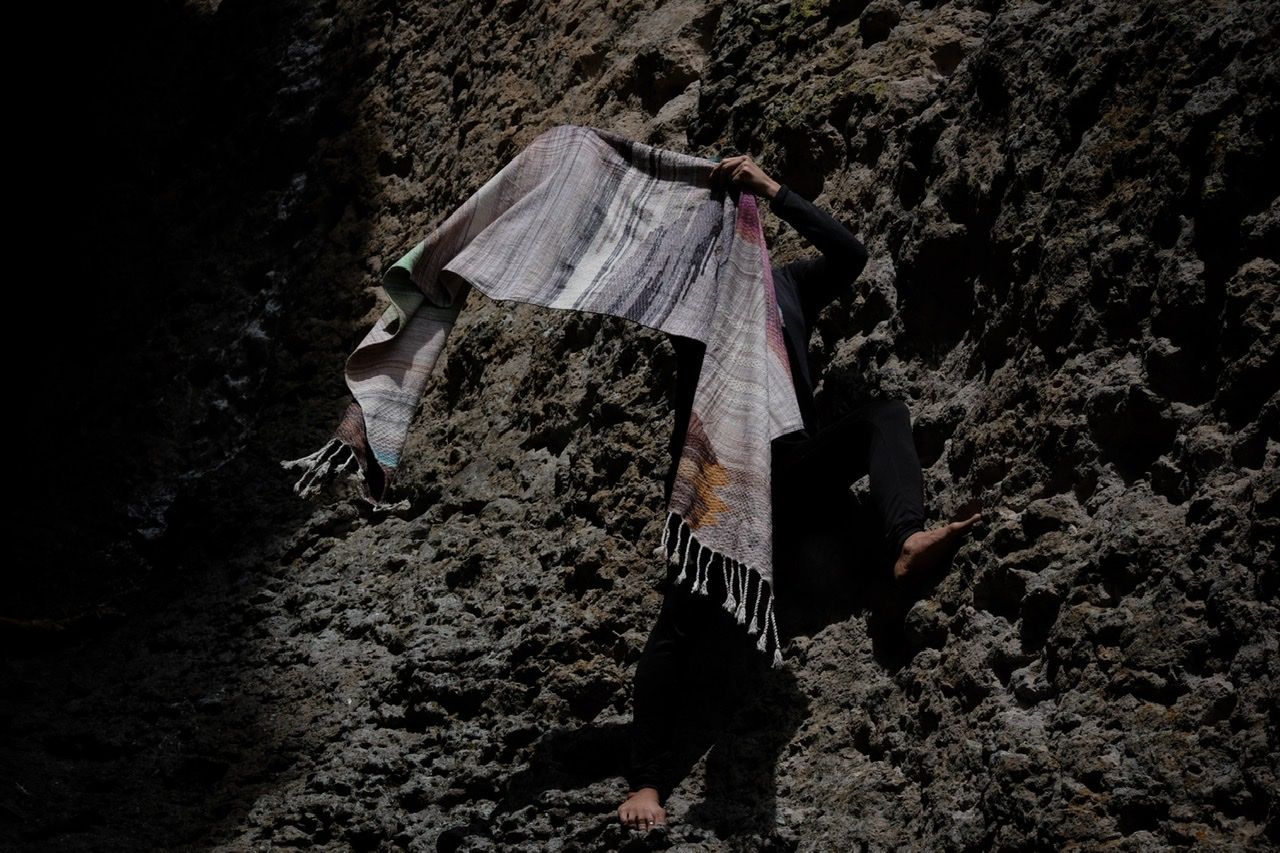 A woman wears a handwoven, diamond pattern shawl in blues, grey, black, brown, pink and green while standing in a stone canyon