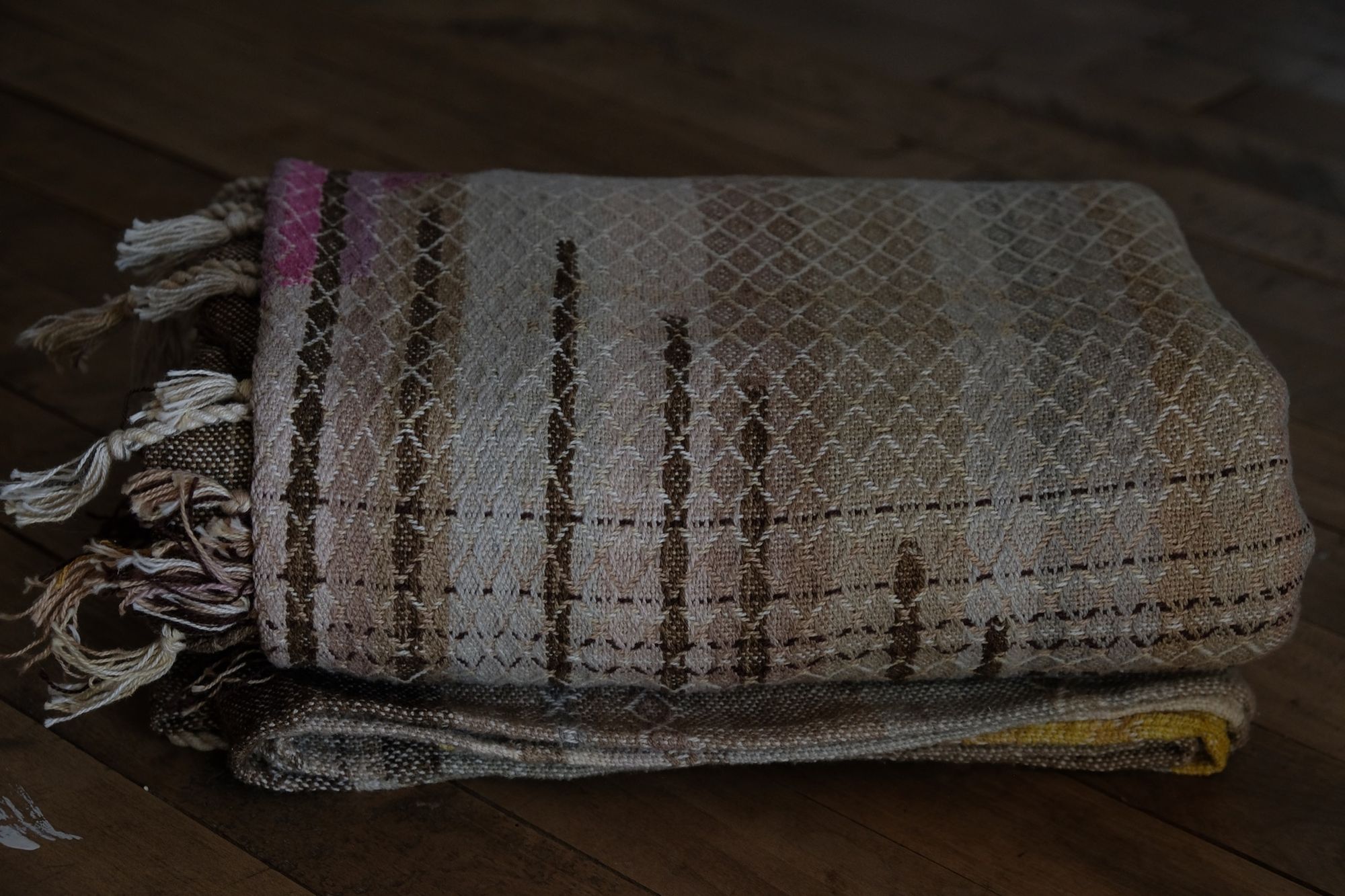 Handwoven fabric in soft shades of grey, pink, yellow and earth laying on a wooden floor