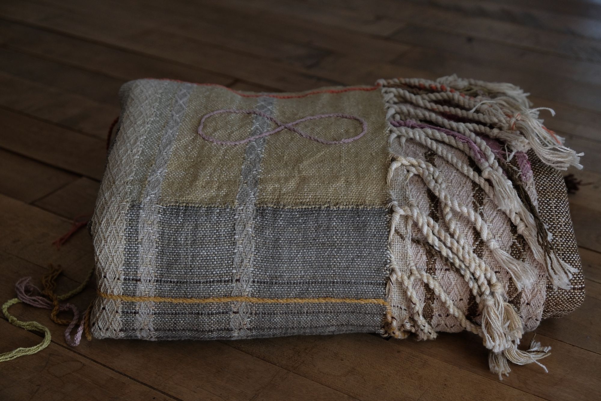 Handwoven fabric in soft shades of grey, pink, yellow and earth laying on a wooden floor