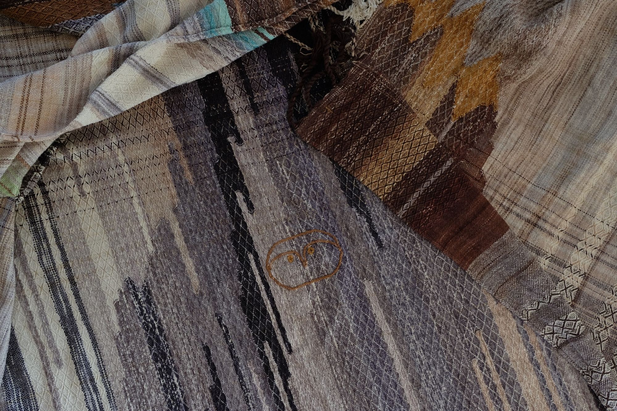 A handwoven, diamond pattern shawl in blues, grey, black, brown, pink and green laying on a wooden flor