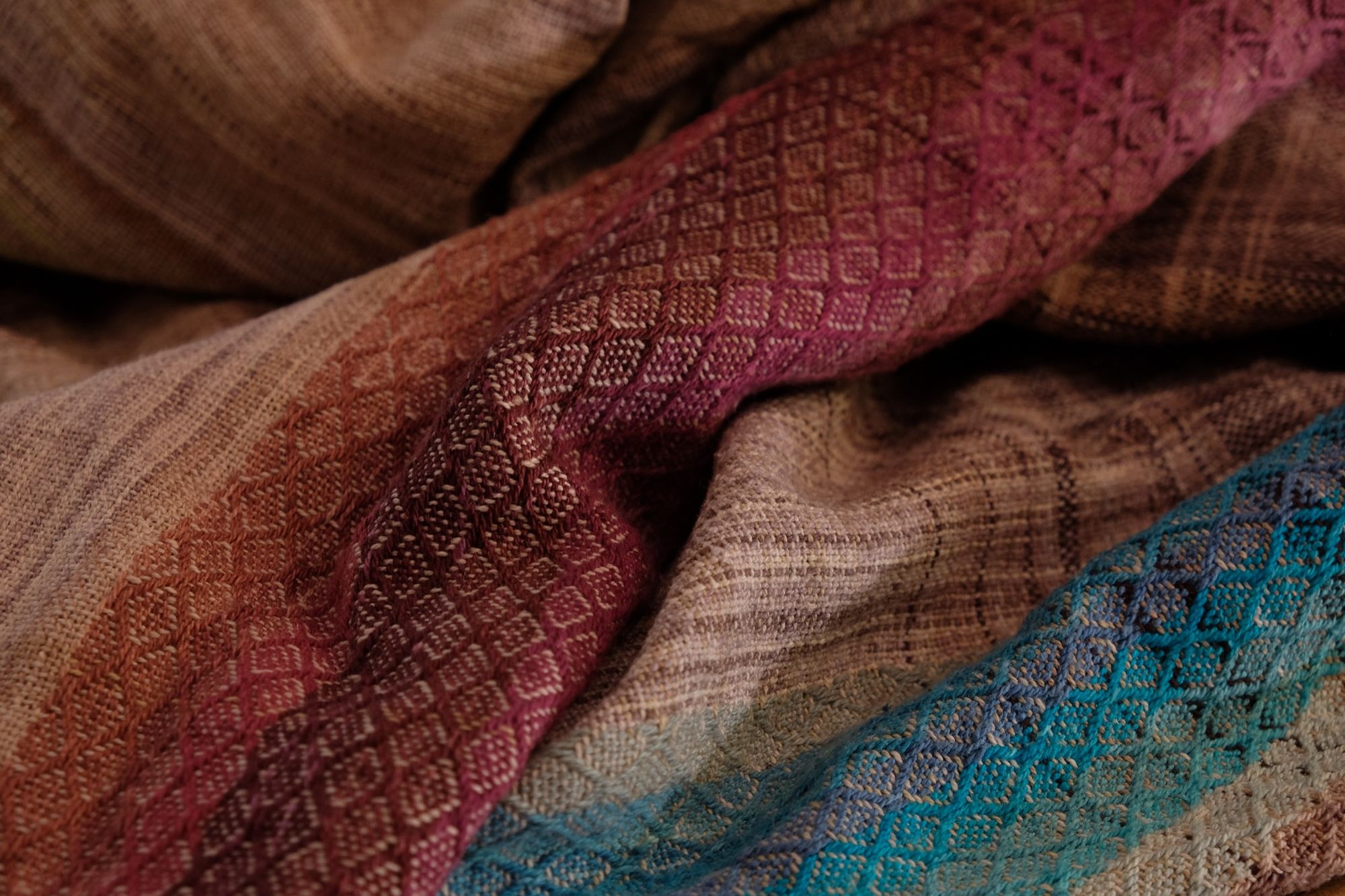A handwoven, diamond pattern shawl in blues, grey, black, brown, pink and green laying on a wooden flor