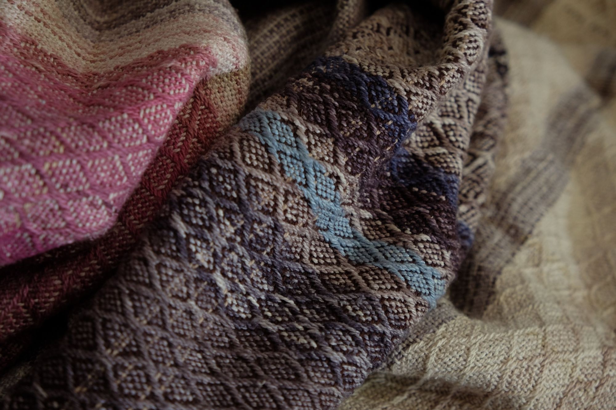 A handwoven, diamond pattern shawl in blues, grey, black, brown, pink and green laying on a wooden flor