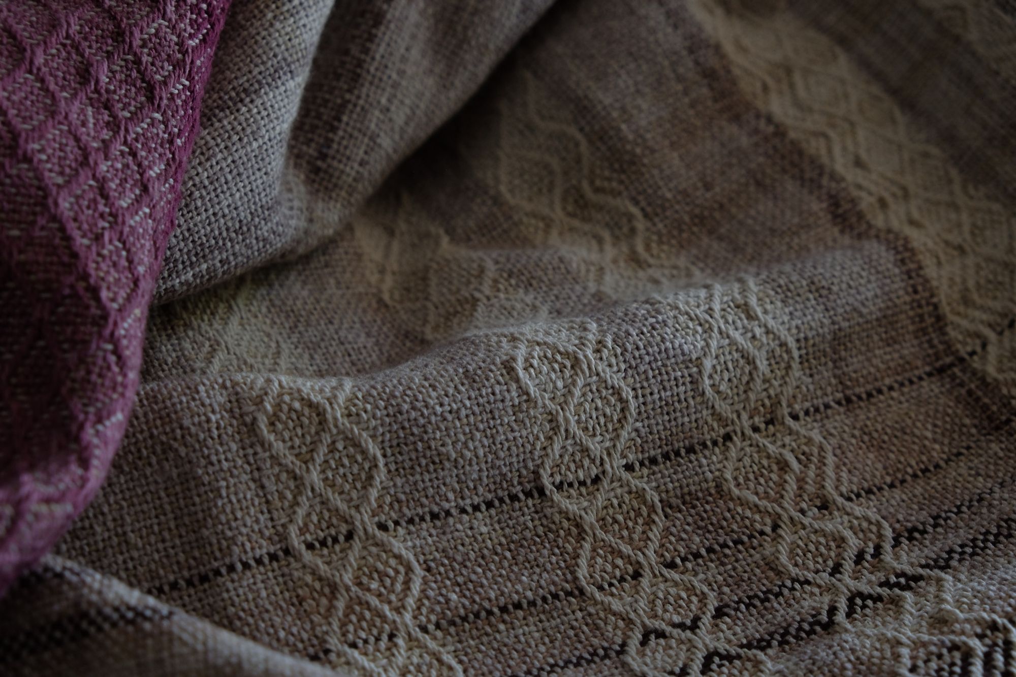 A handwoven, diamond pattern shawl in blues, grey, black, brown, pink and green laying on a wooden flor