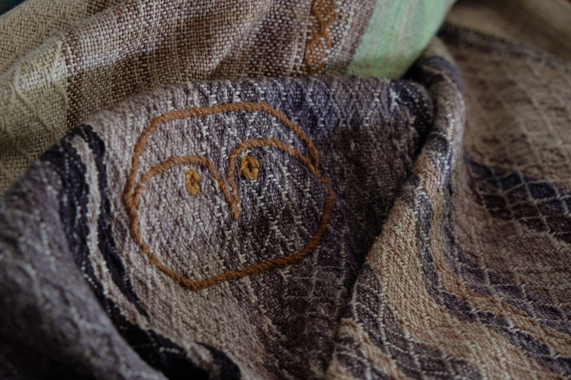 a small owl detail on A handwoven, diamond pattern shawl in blues, grey, black, brown, pink and green laying on a wooden floor