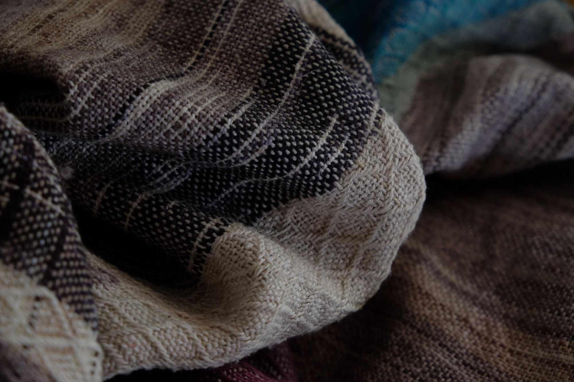 A handwoven, diamond pattern shawl in blues, grey, black, brown, pink and green laying on a wooden floor