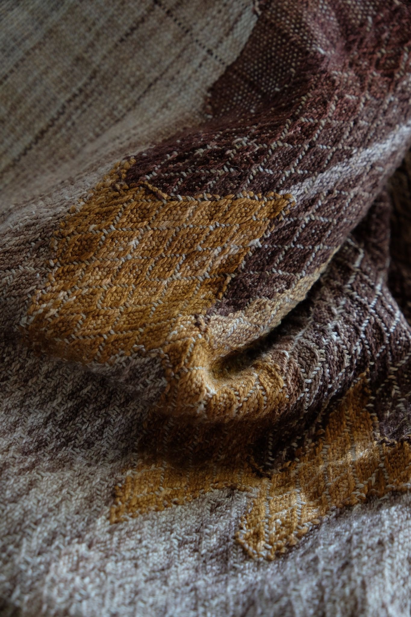 A handwoven, diamond pattern shawl in blues, grey, black, brown, pink and green laying on a wooden floor