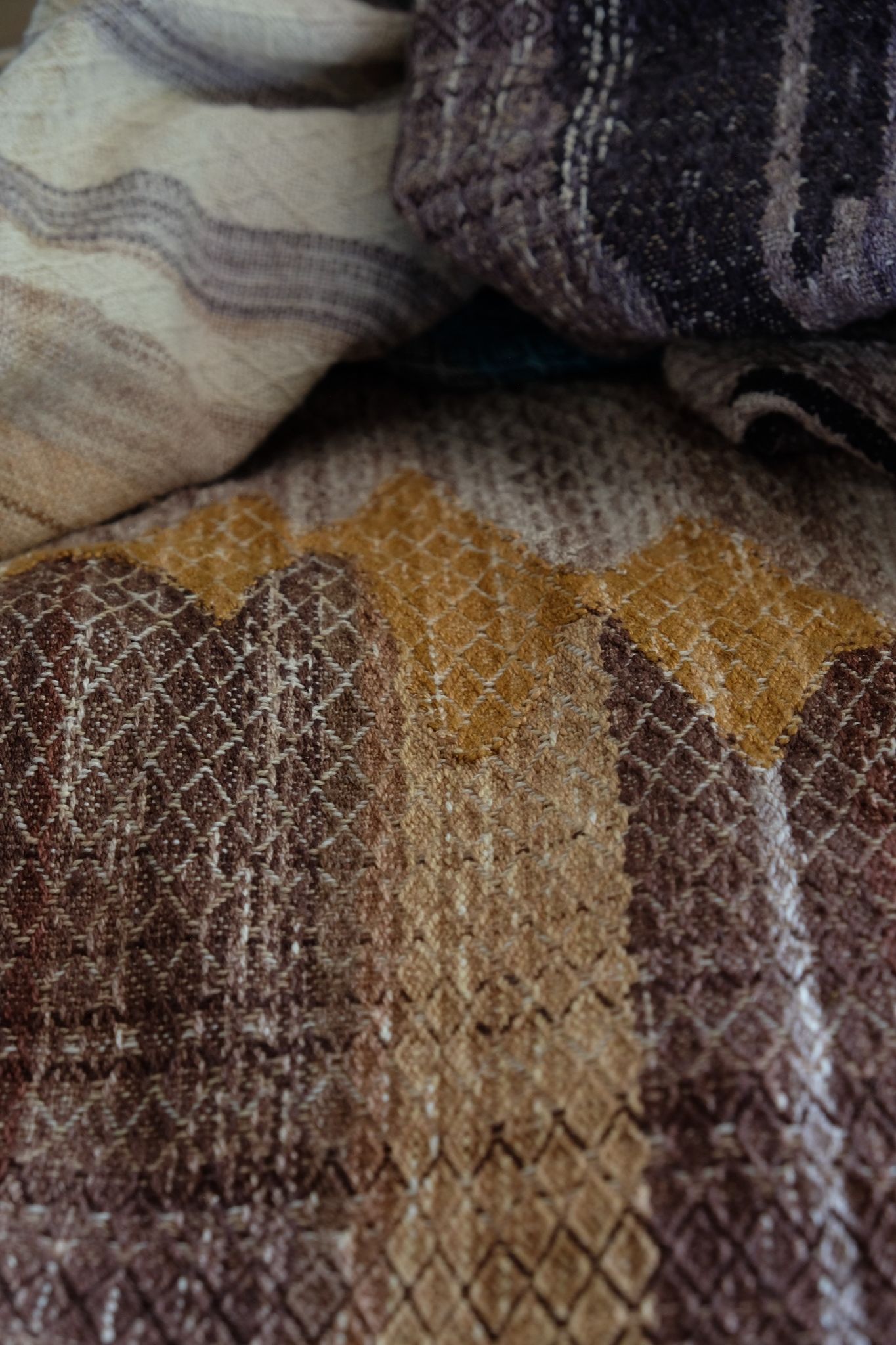 A handwoven, diamond pattern shawl in blues, grey, black, brown, pink and green laying on a wooden floor