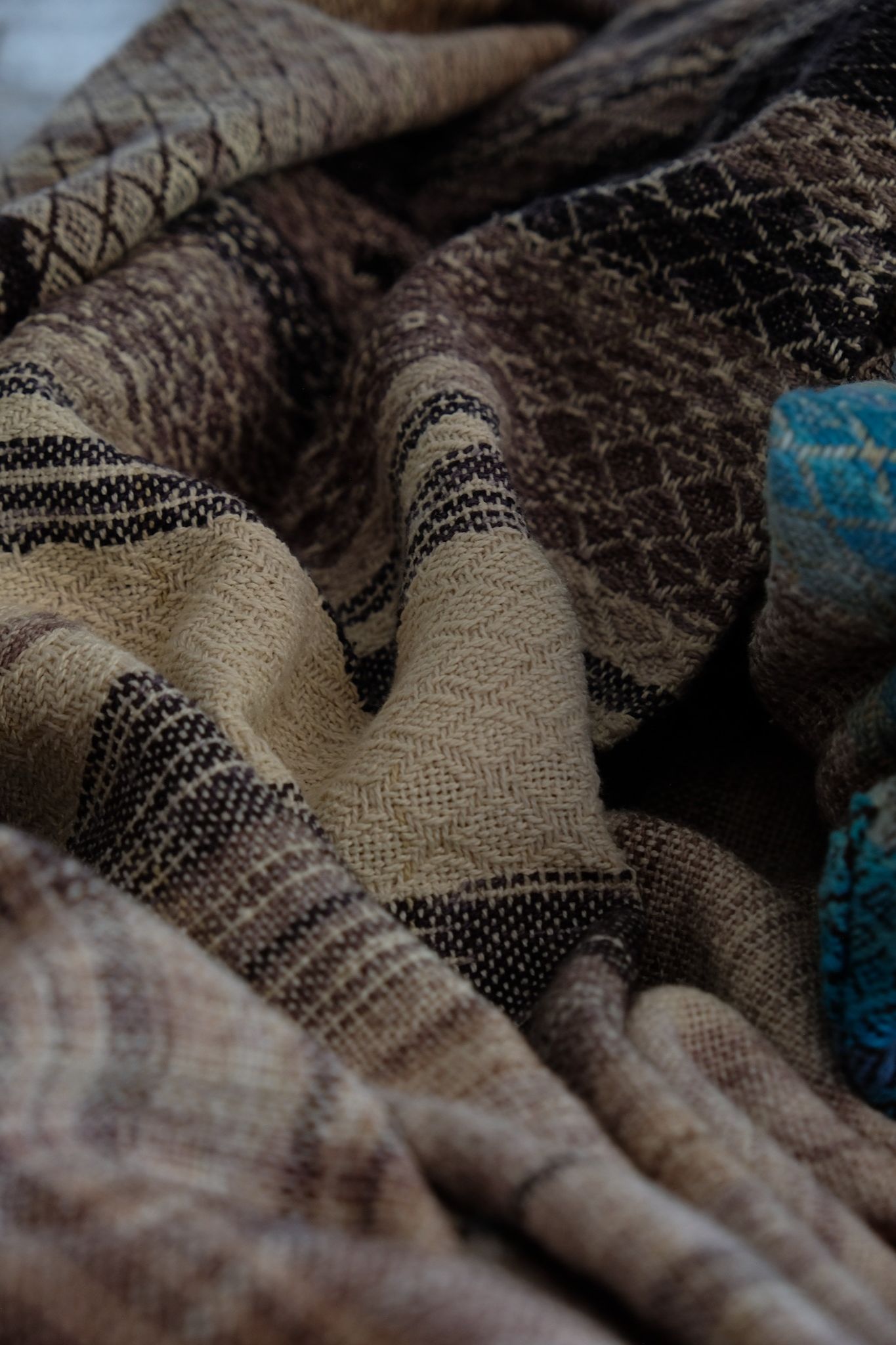 A handwoven, diamond pattern shawl in blues, grey, black, brown, pink and green laying on a wooden floor