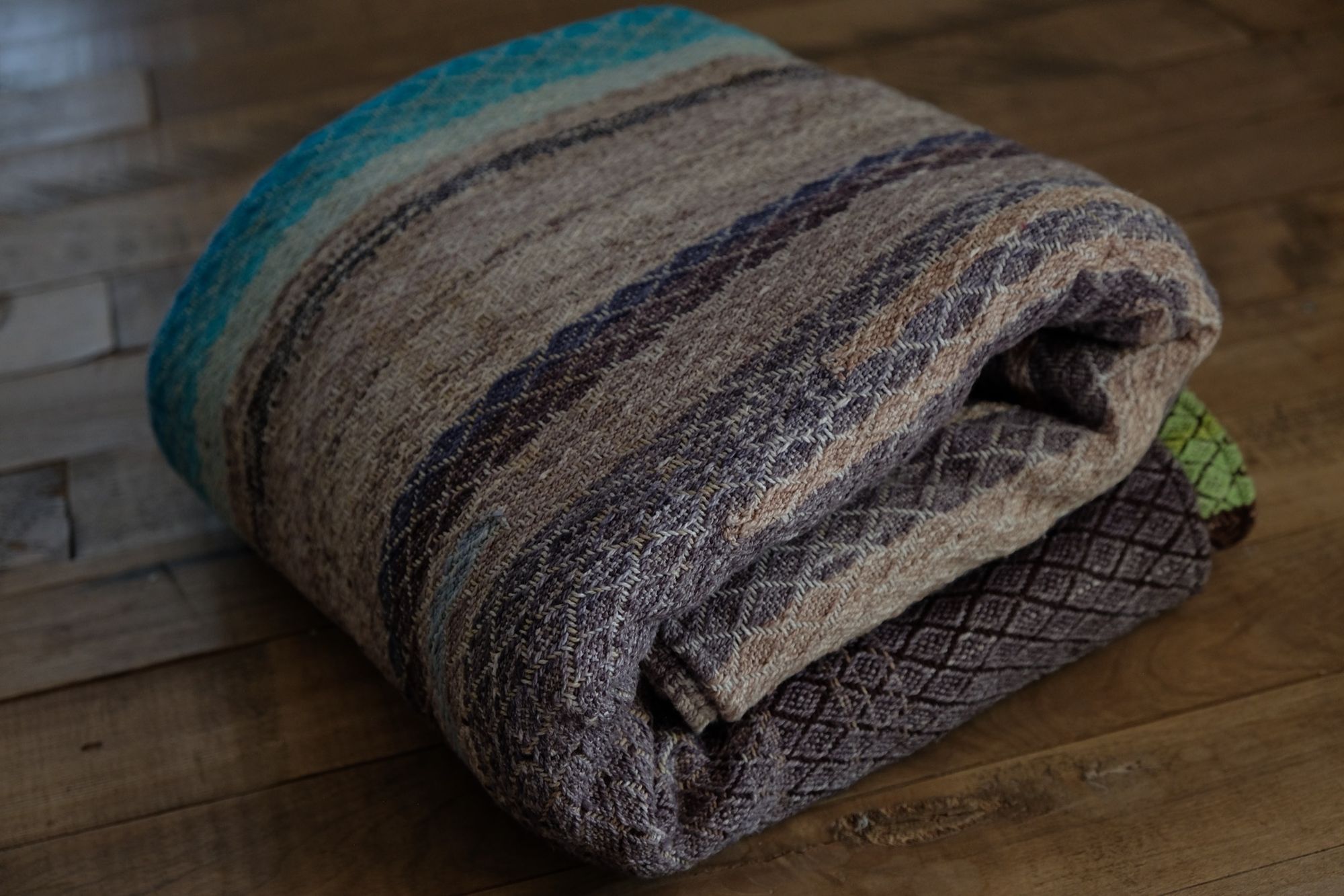 A handwoven, diamond pattern shawl in blues, grey, black, brown, pink and green laying on a wooden floor