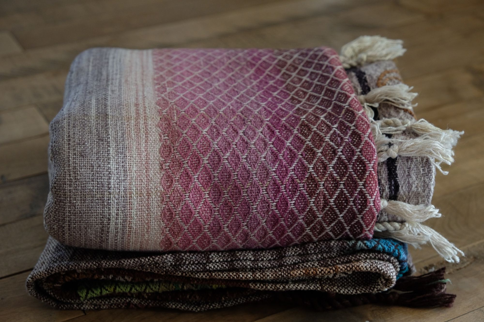 A handwoven, diamond pattern shawl in blues, grey, black, brown, pink and green laying on a wooden floor