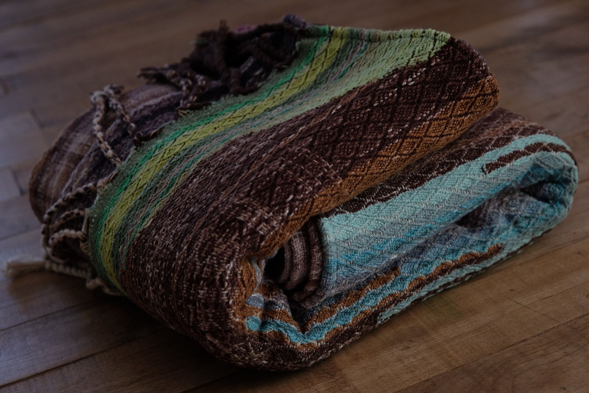 A handwoven, diamond pattern shawl in blues, grey, black, brown, pink and green laying on a wooden floor
