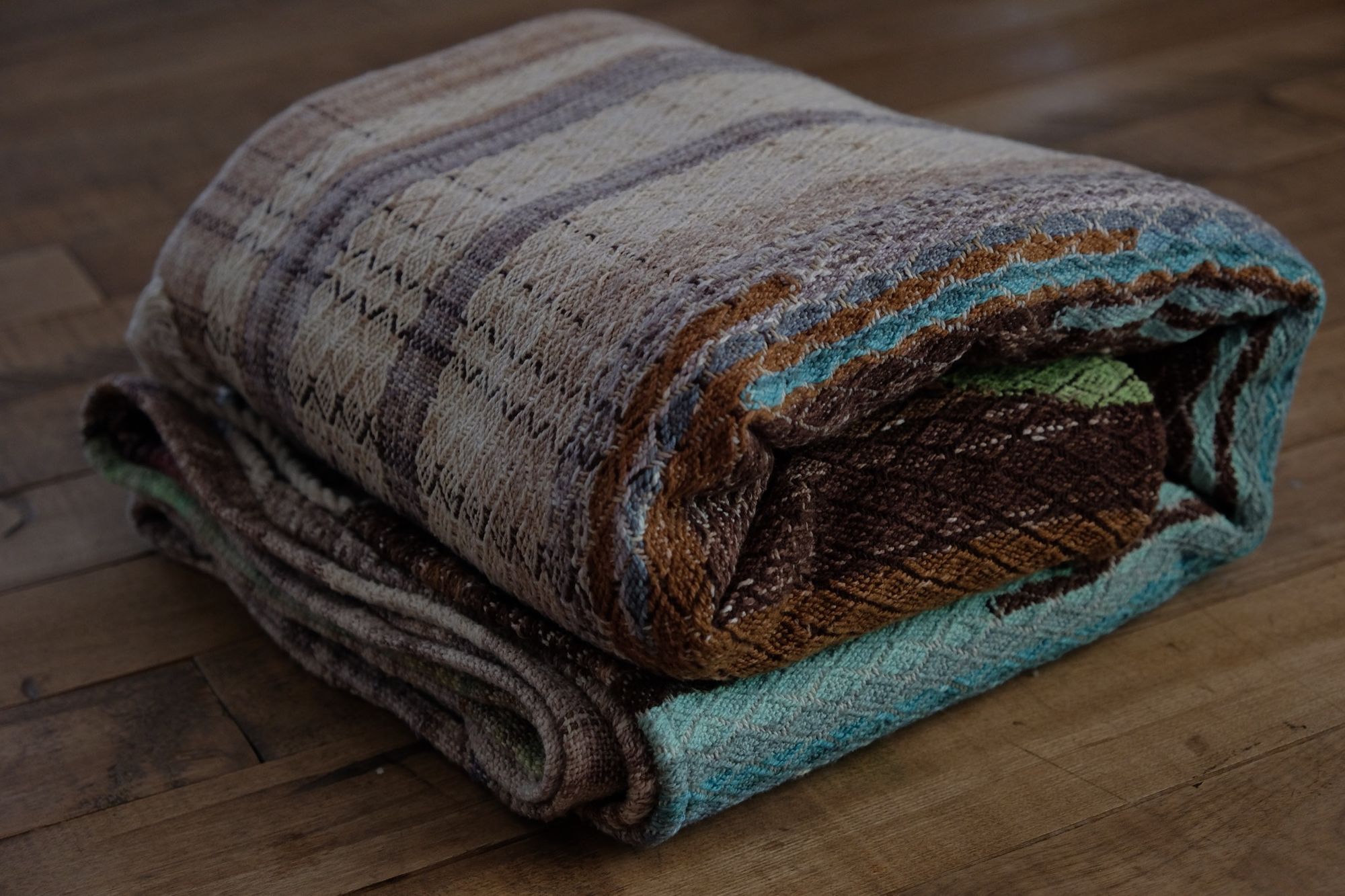 A handwoven, diamond pattern shawl in blues, grey, black, brown, pink and green laying on a wooden floor