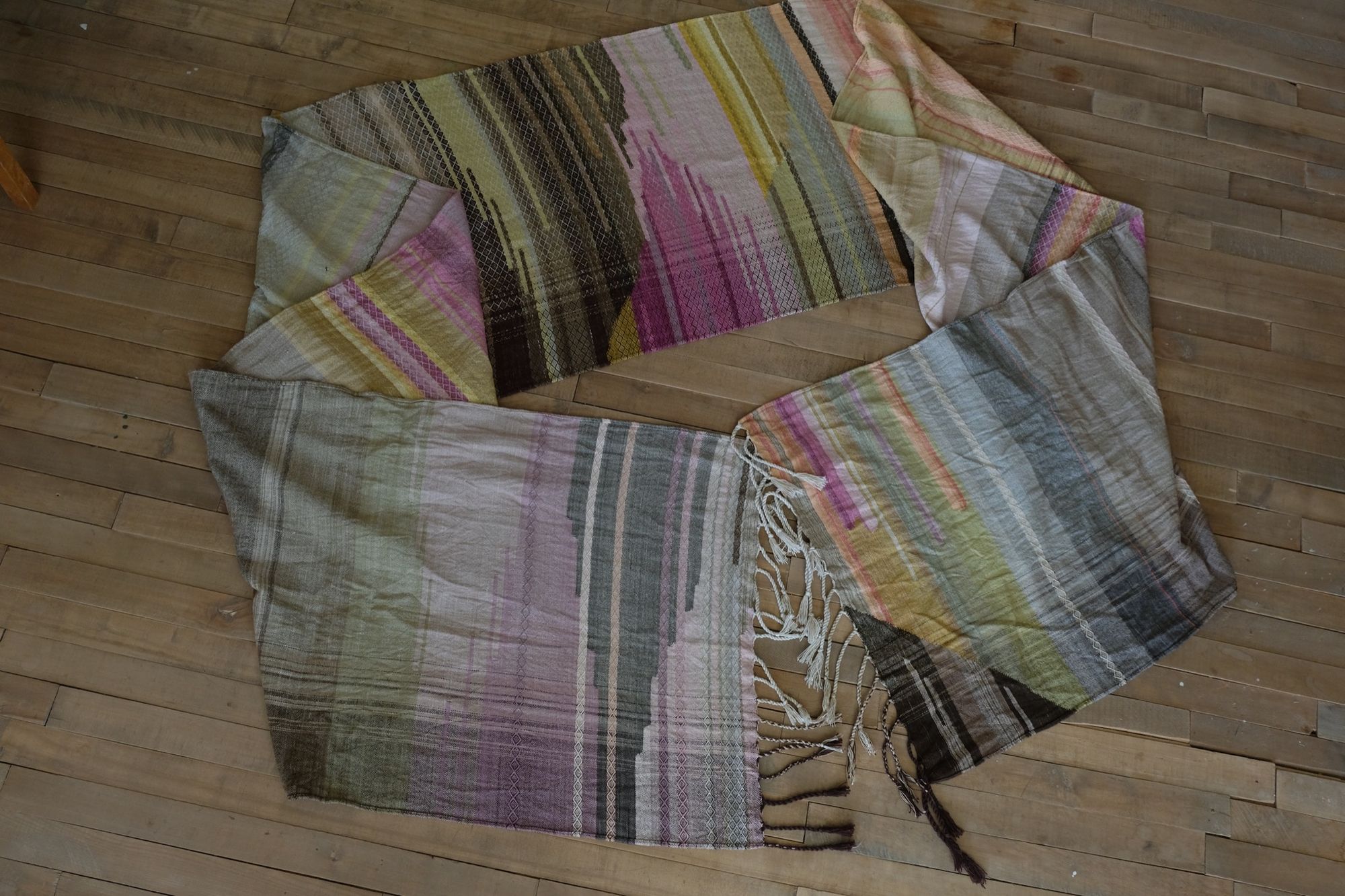 A long length of handwoven fabric with a diamond pattern in a naturally dyed rainbow of color, brown, grey, pink, purple, yellow, green, orange and lavender lays on a wooden floor