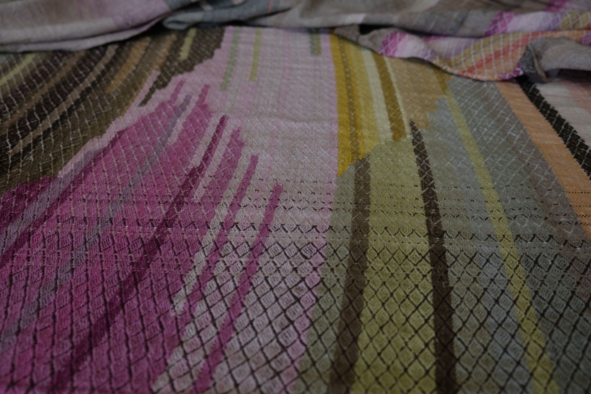 detail of A long length of handwoven fabric with a diamond pattern in a naturally dyed rainbow of color, brown, grey, pink, purple, yellow, green, orange and lavender lays on a wooden floor