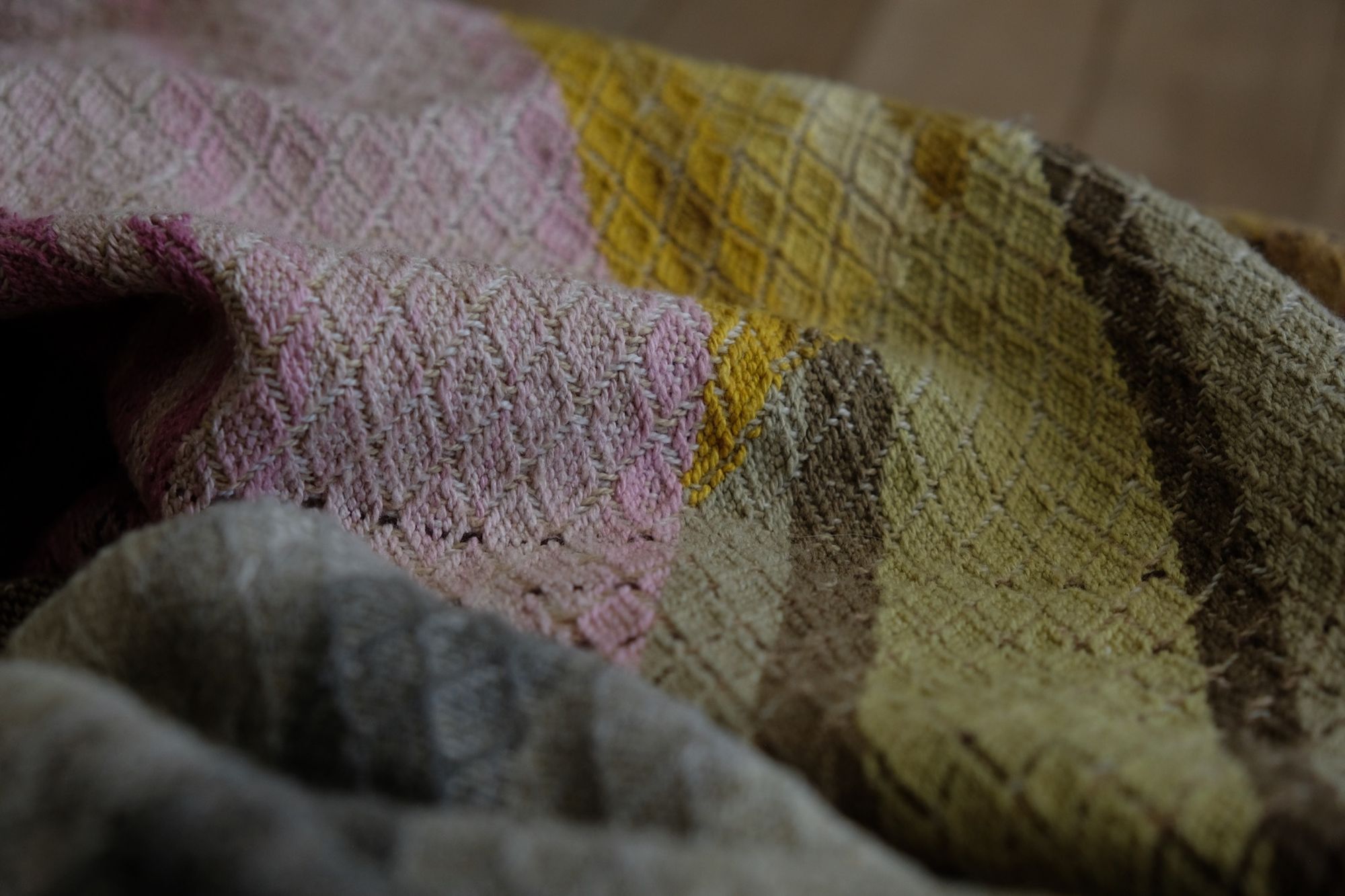 detail of A long length of handwoven fabric with a diamond pattern in a naturally dyed rainbow of color, brown, grey, pink, purple, yellow, green, orange and lavender lays on a wooden floor