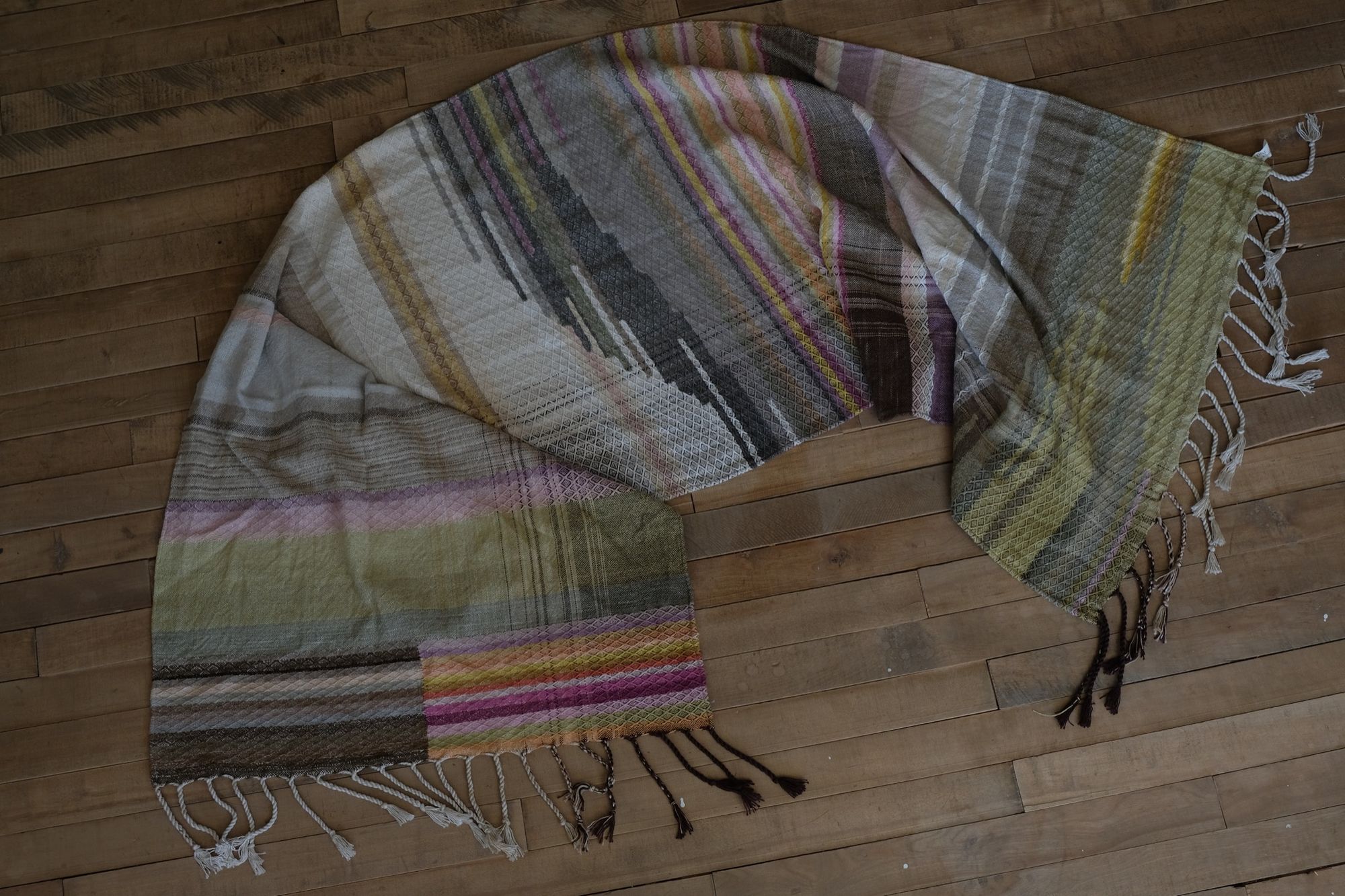 Handwoven fabric in a naturally dyed rainbow of grey, browns, greens, pinks, purples, yellow and orange lays on a wooden floor