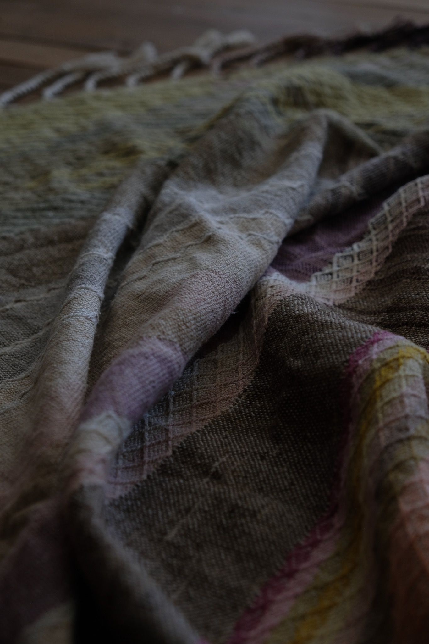 Detail of Handwoven fabric in a naturally dyed rainbow of grey, browns, greens, pinks, purples, yellow and orange lays on a wooden floor
