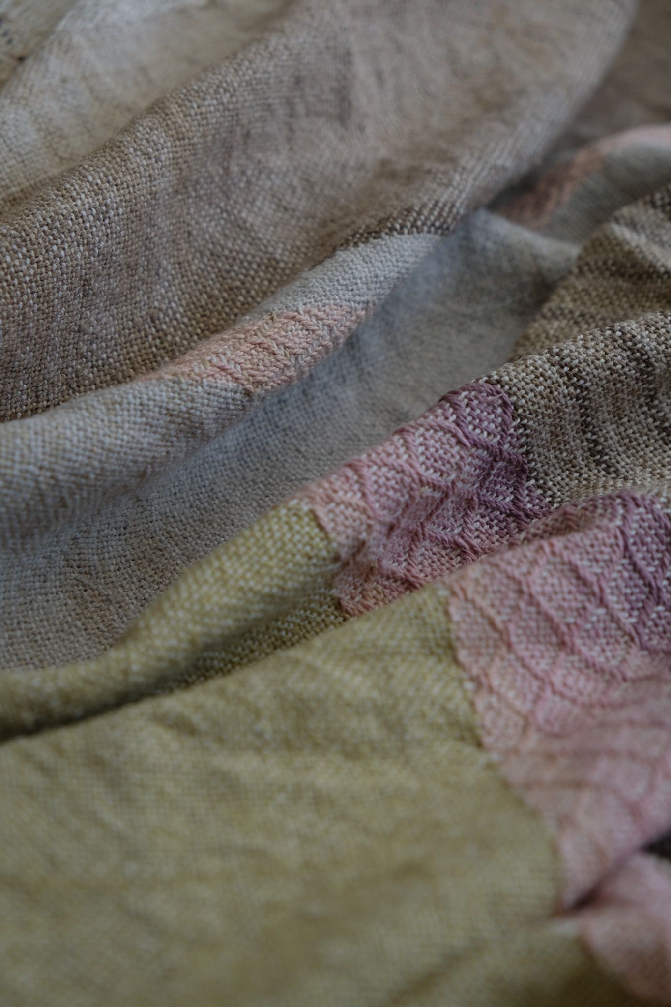 Detail of Handwoven fabric in a naturally dyed rainbow of grey, browns, greens, pinks, purples, yellow and orange lays on a wooden floor
