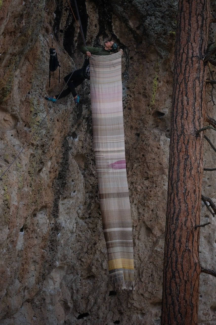 A woman hangs from ropes off of a red, black and tan cliff while holding a large piece of handwoven fabric