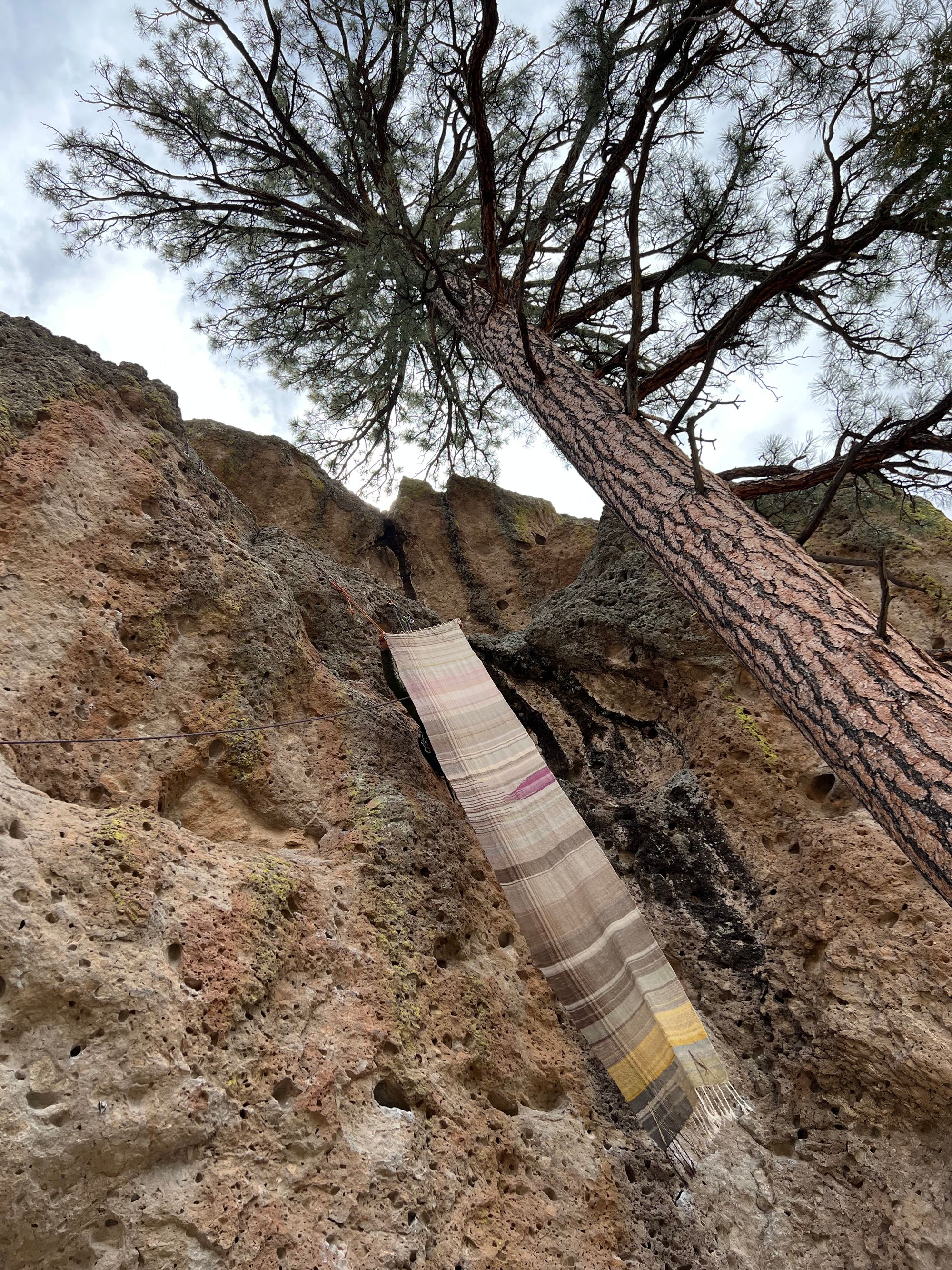 A woman hangs from ropes off of a red, black and tan cliff while holding a large piece of handwoven fabric