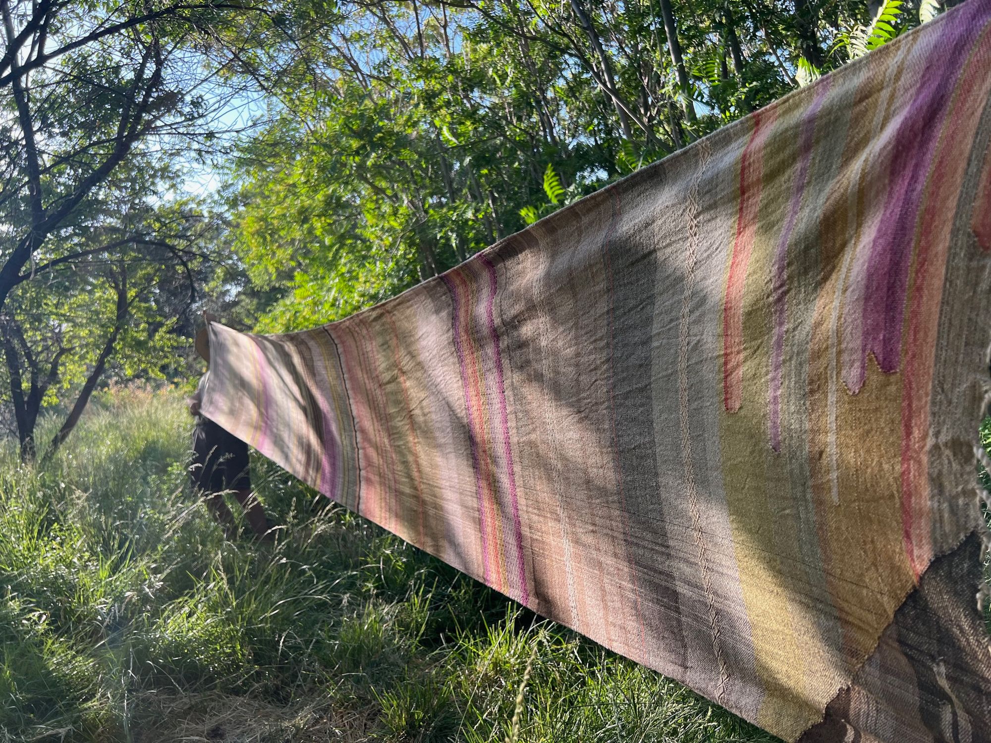 a long piece of handwoven fabric in a naturally dyed rainbow is stretched out in a dense green forest
