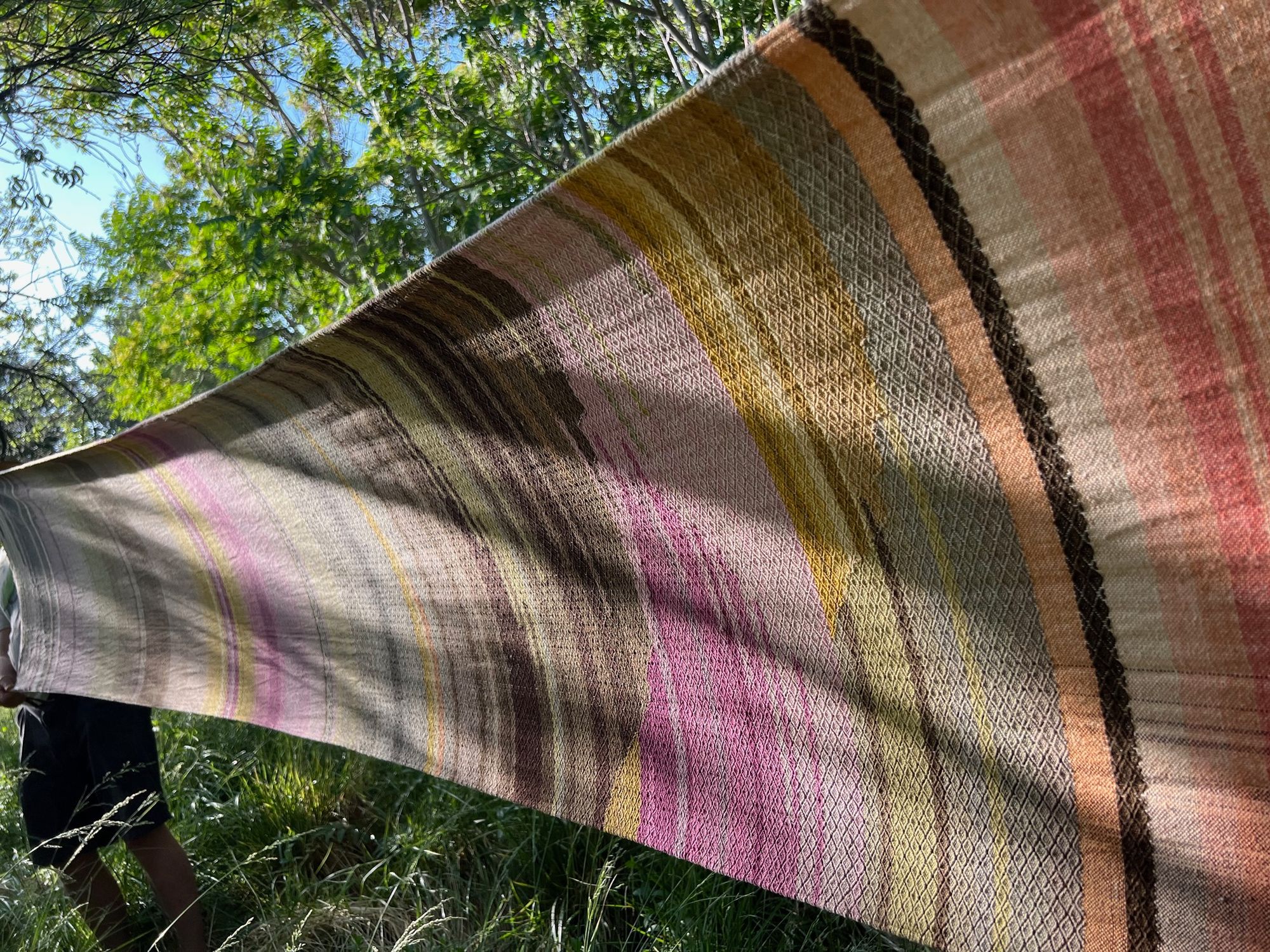 a long piece of handwoven fabric in pink, brown, purple, green, yellow, grey and grey is stretched out in a dense green forestdetail of A long length of handwoven fabric with a diamond pattern in a naturally dyed rainbow of color, brown, grey, pink, purple, yellow, green, orange and lavender lays on a wooden floor
