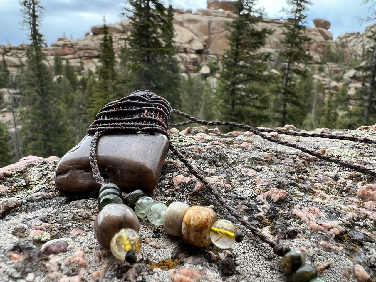 A small brown knotwork basket with beads and petrified wood hangs from a corded necklace on a lichen covered rock