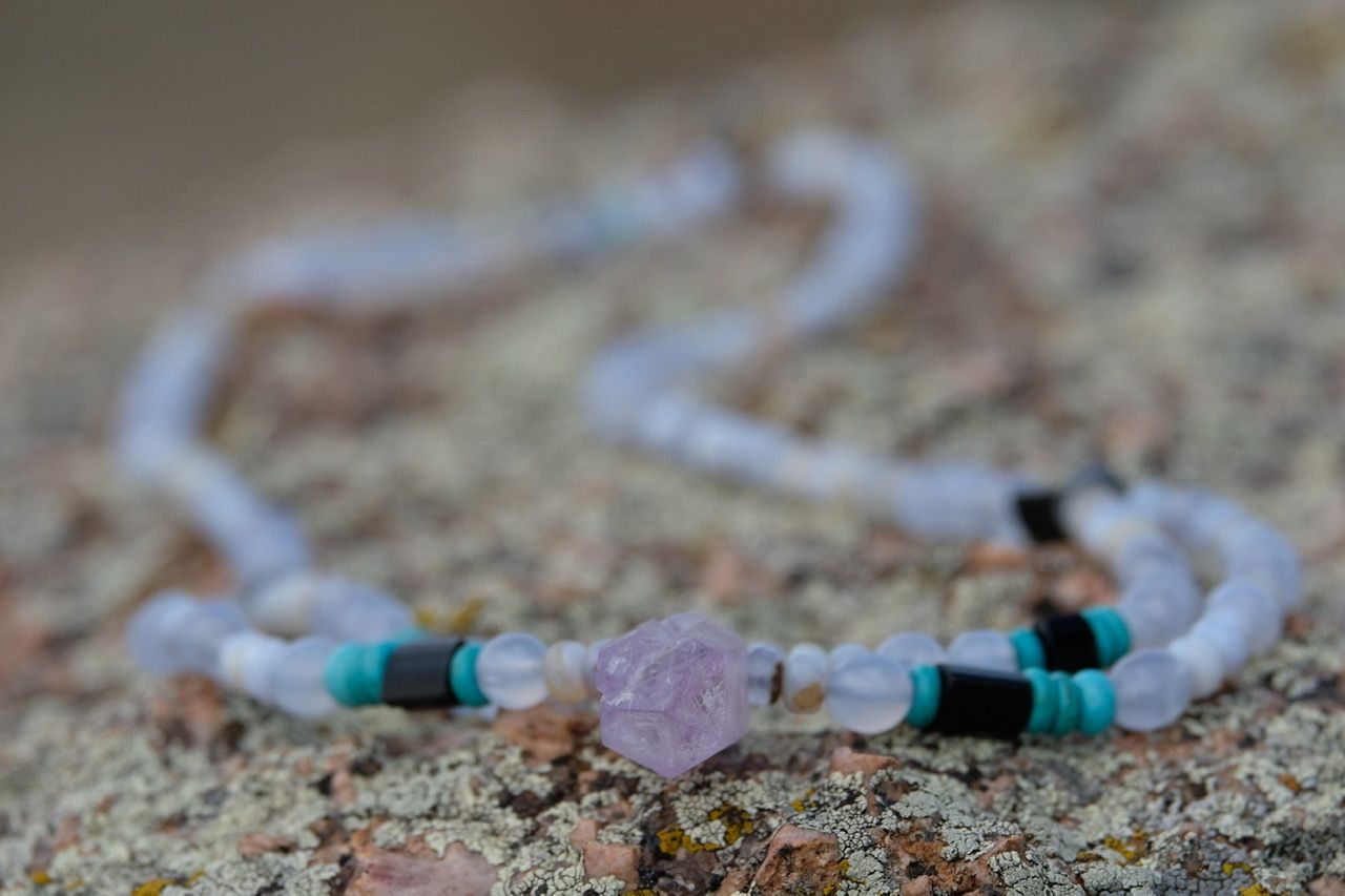 A purple amethyst, blue chalk, turquoise and black tourmaline necklace lays on a lichen covered rock
