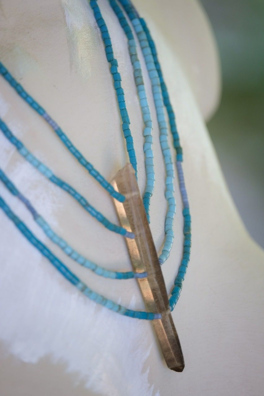 A 4 strand pale blue beaded necklace with a thin crystal centerpiece is on a white painted mannequin 