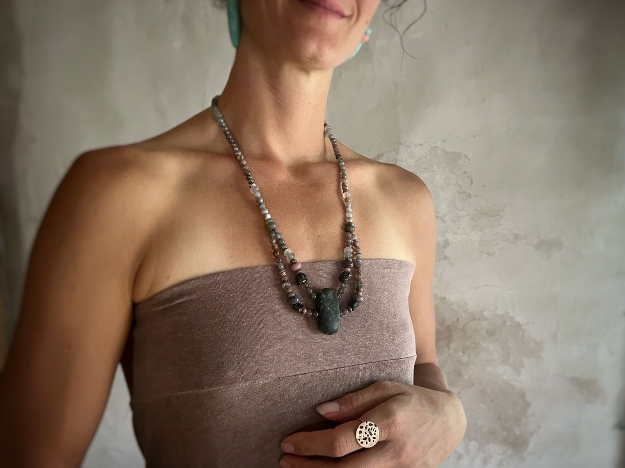 A woman wearing a pale purple top and a long stone necklace stands in front of a white mud wall