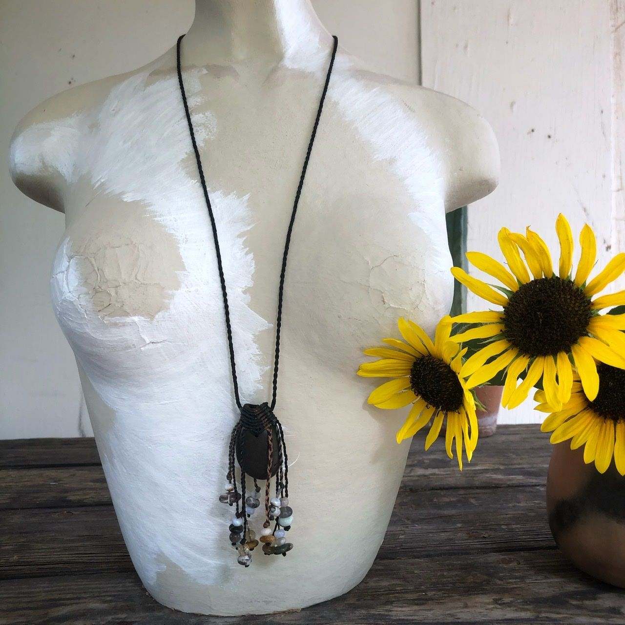 A smooth black ocean tumbled stone talisman necklace with braided dangling beads, rests on a white mannequin on a wood table with sunflowers in the background 