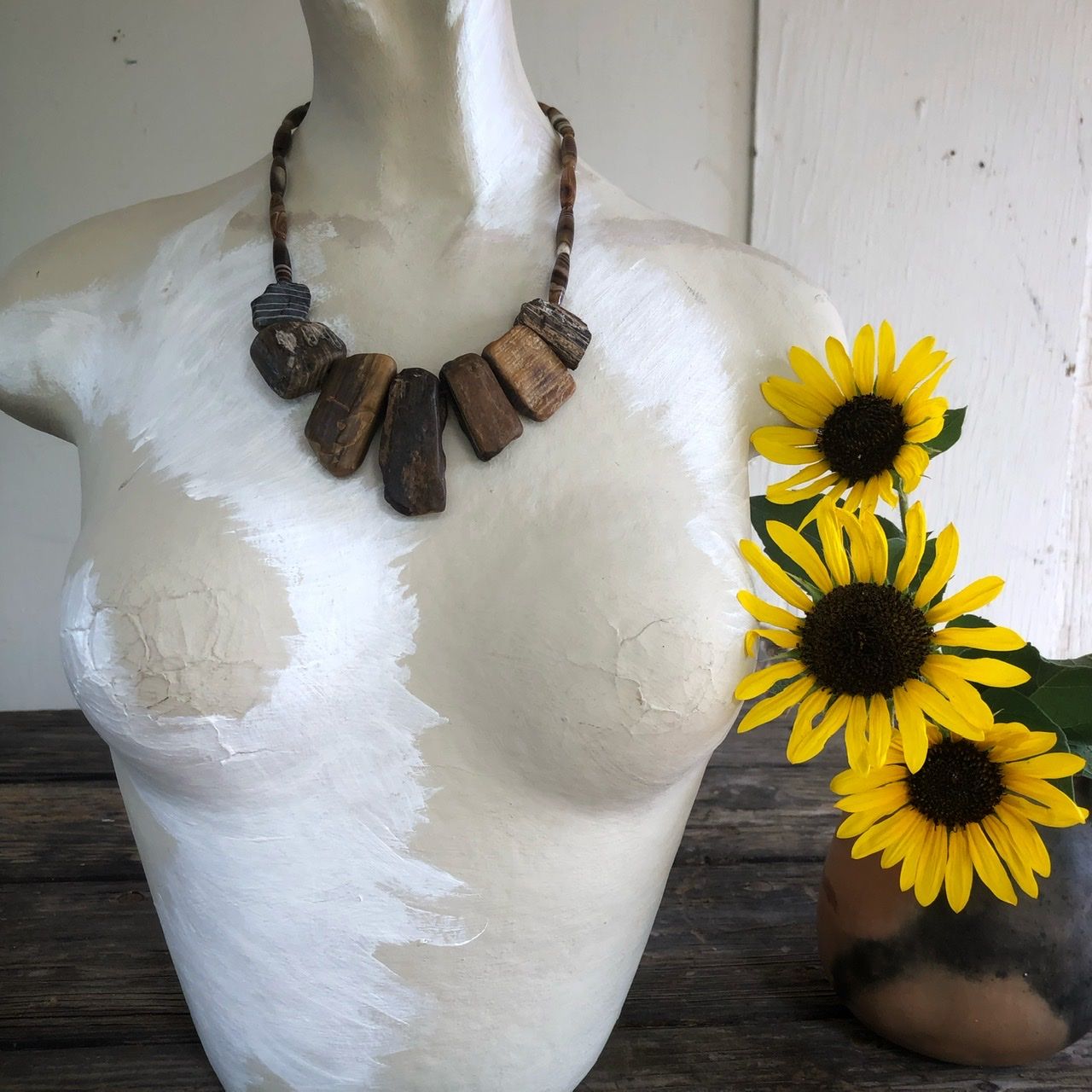 A petrified wood and banded agate necklace rests on a white mannequin won a wood table with sunflowers in the background as