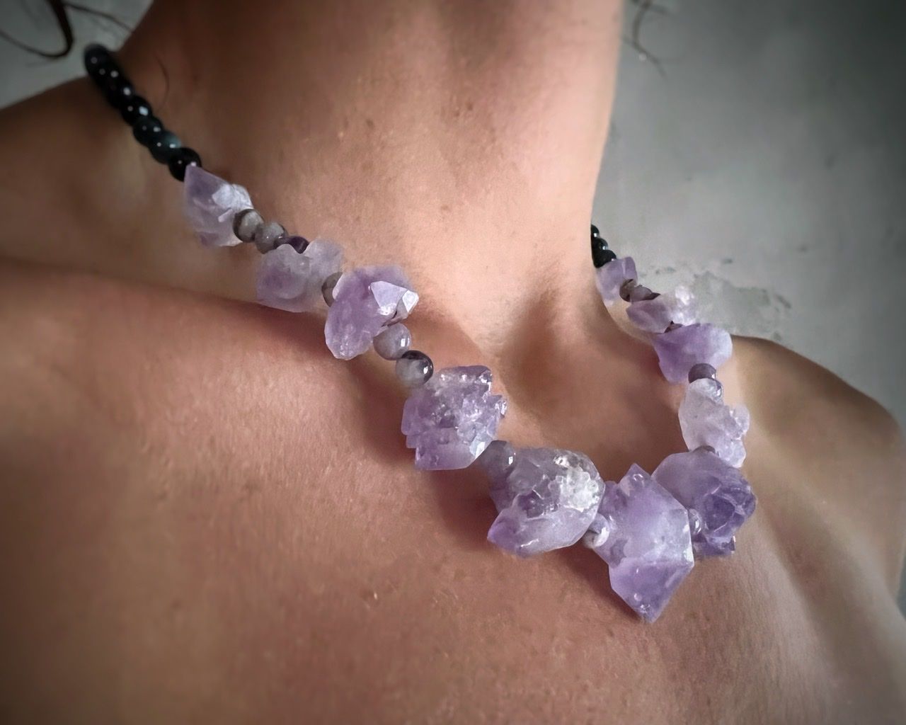 A woman wearing a pale purple top and a short amethyst crystal necklace stands in front of a white mud wall