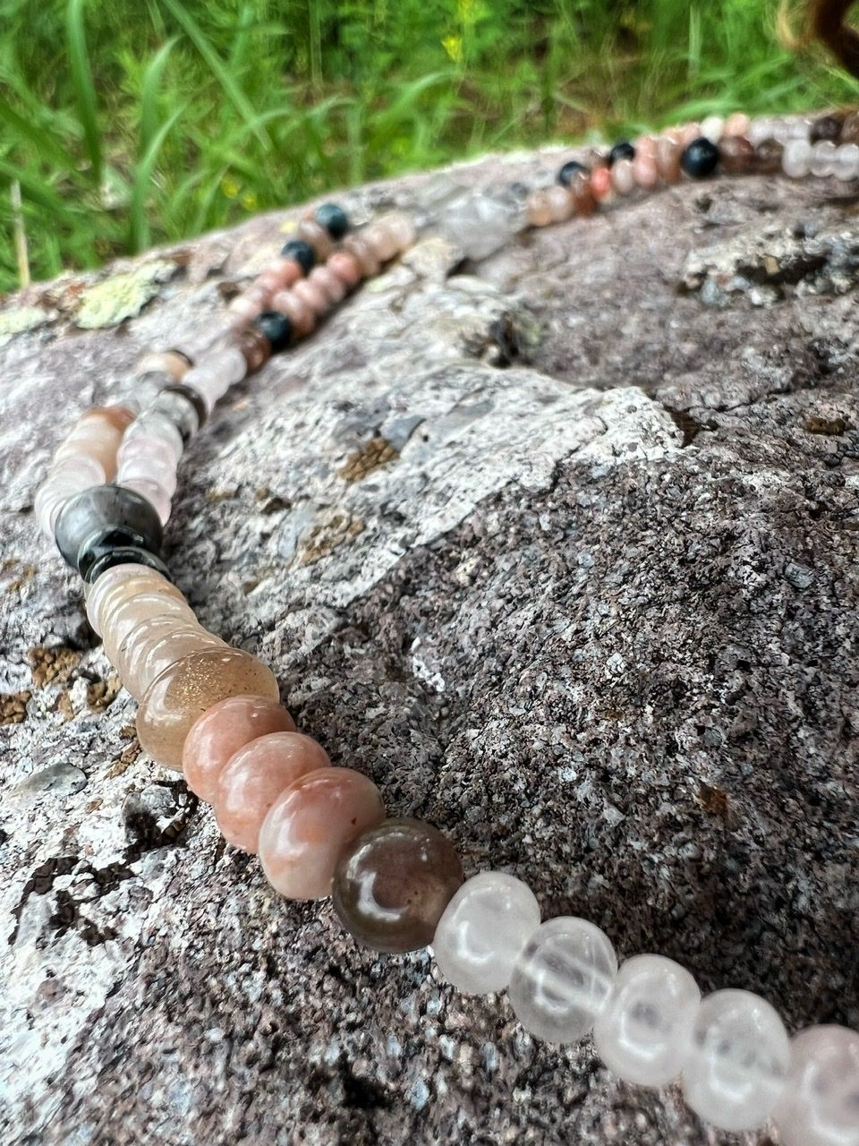 a stone beaded necklace in pale pinks, dusty pinks, black and grays lays on a lichen covered stone
