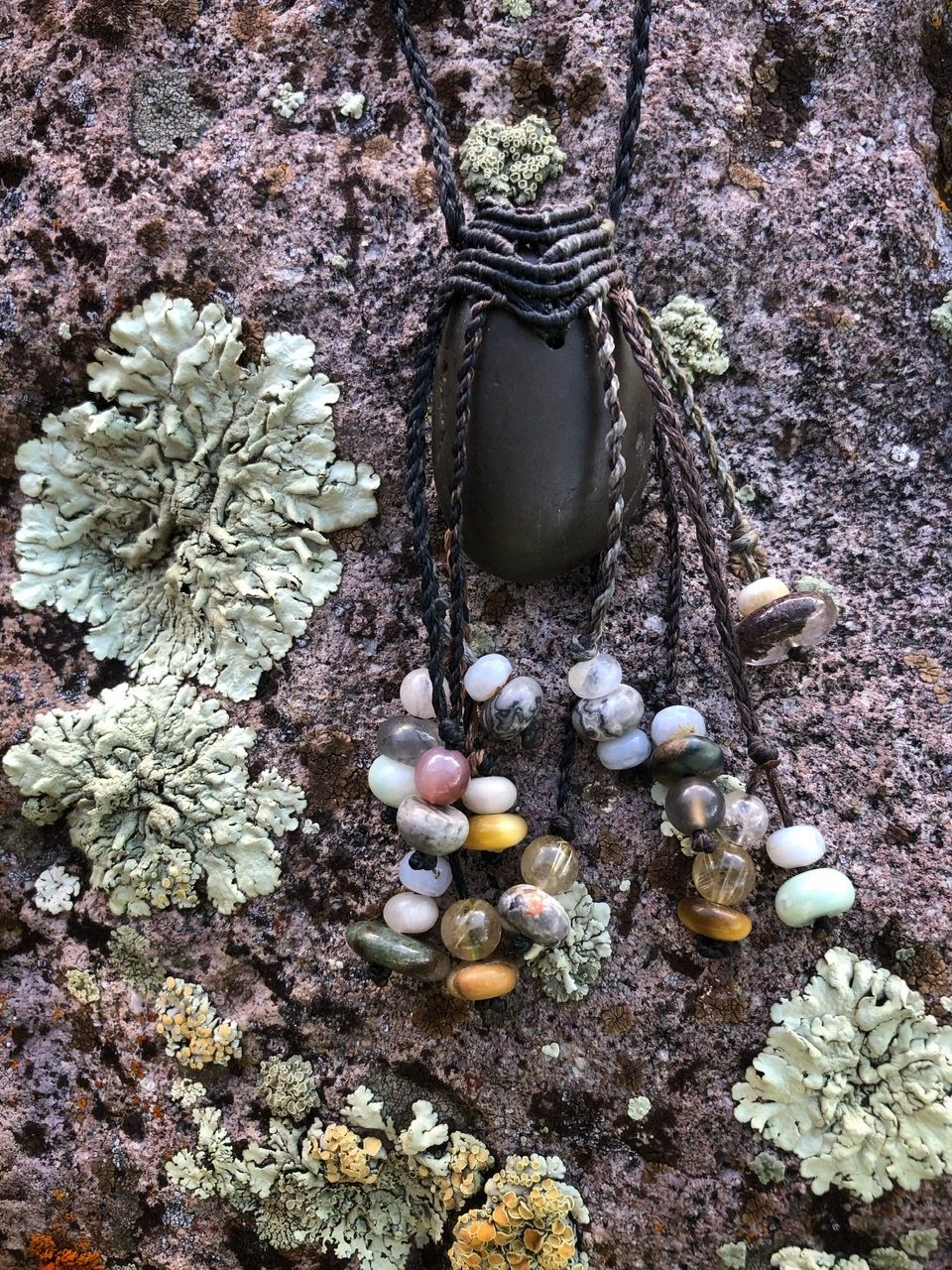 A smooth black ocean tumbled stone talisman necklace with braided dangling beads, rests on a lichen covered Boulder