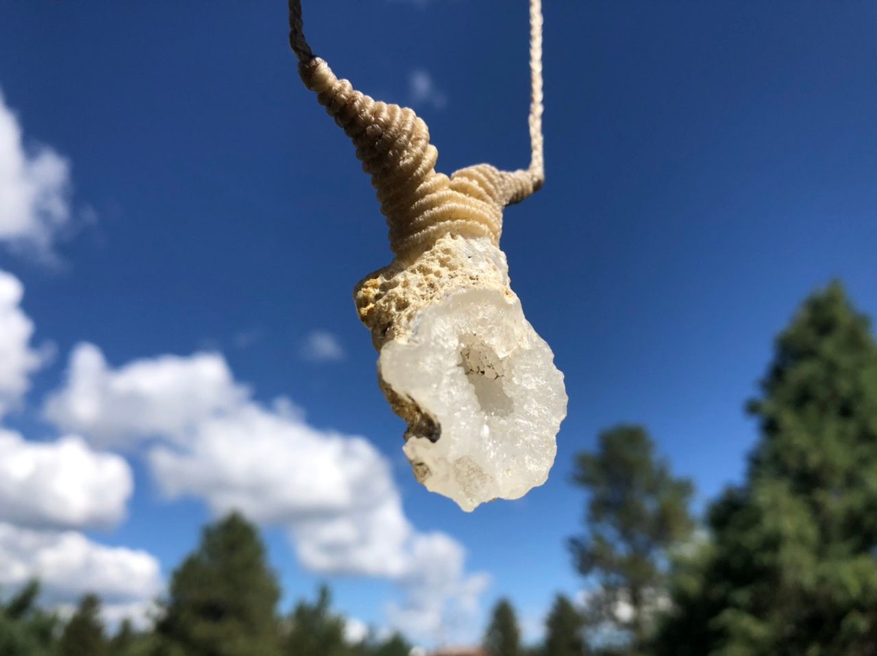  a white thread and crystal necklace Hangs in front of a blue sky and pine trees