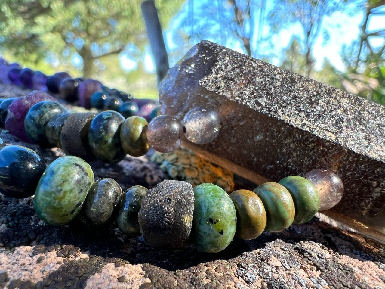 A necklace of purple, green and grey stones and crystals rests on a lichen covered Boulder 
