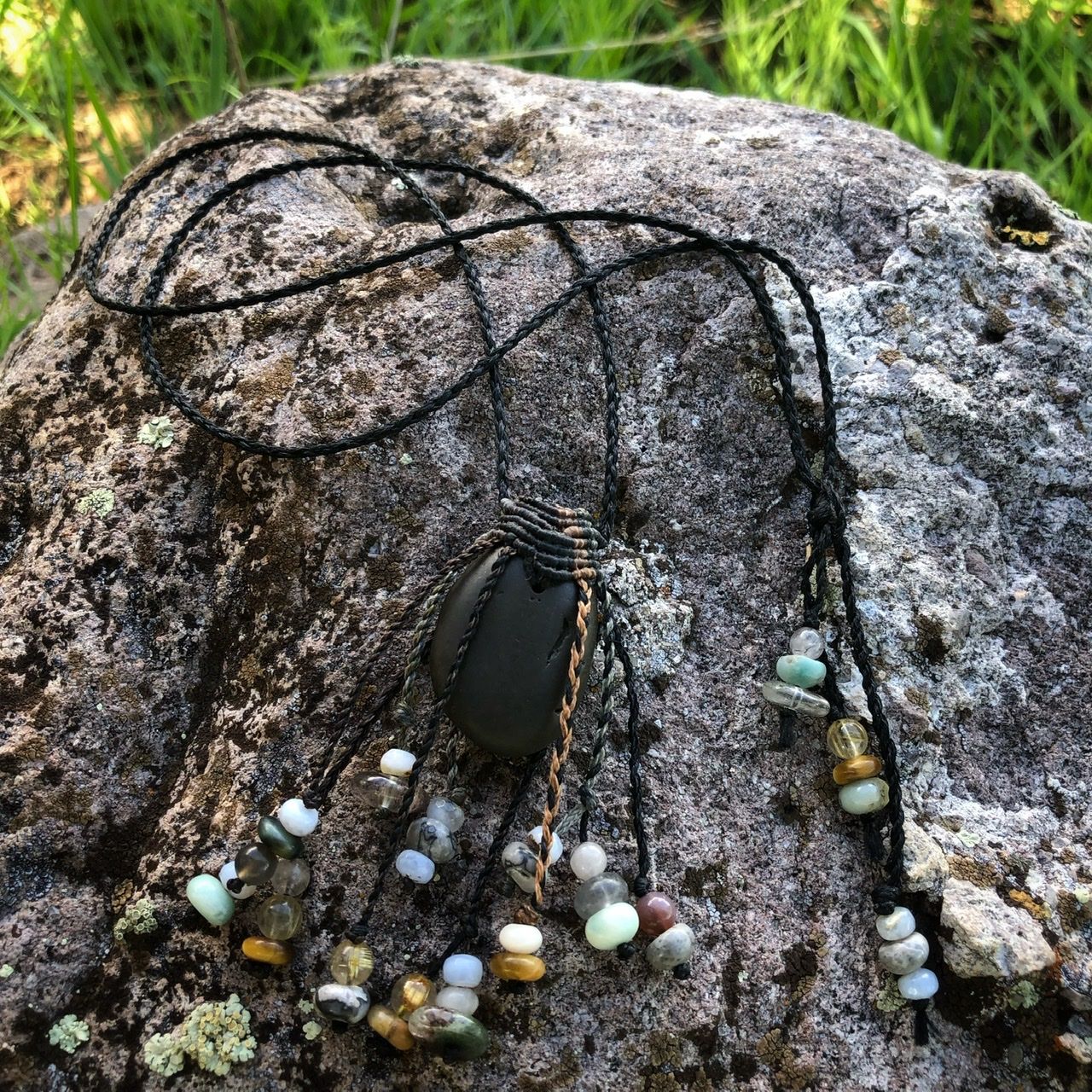 A smooth black ocean tumbled stone talisman necklace with braided dangling beads, rests on a lichen covered Boulder