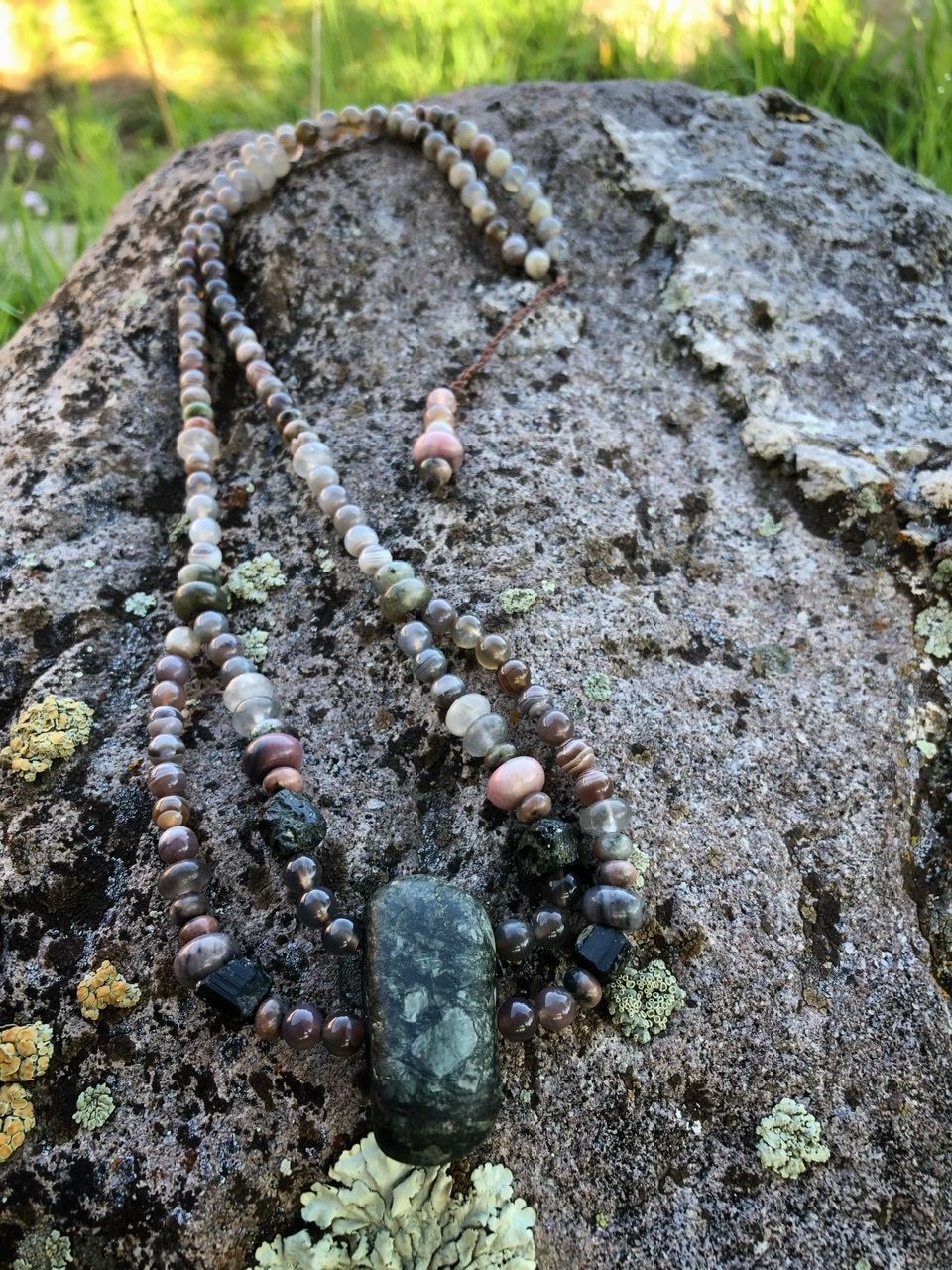 A pink, white, tan and green stone necklace rests on a lichen covered Boulder