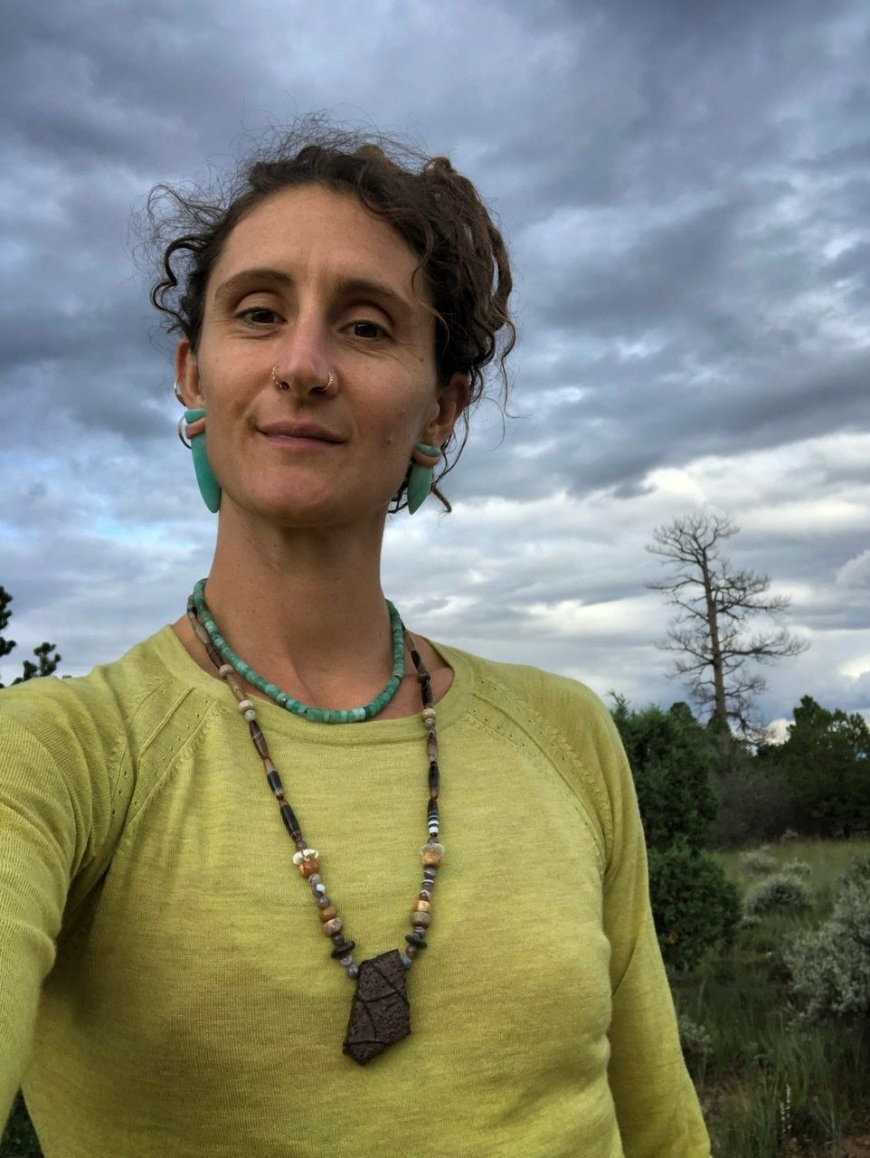 A woman in a chartreuse sweater, green stone earrings and necklace, wears a long earth tone stone Necklace in the forest 