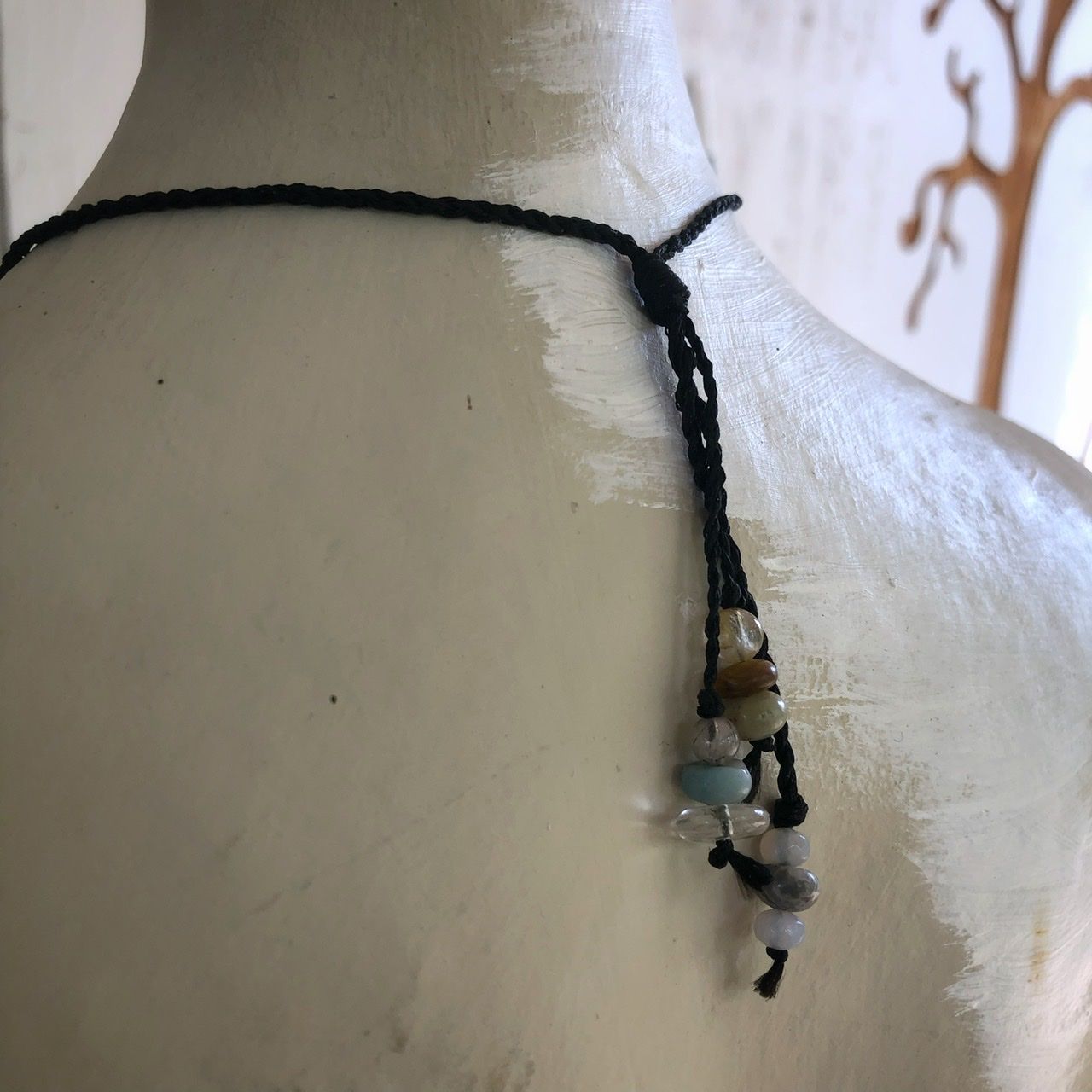 A smooth black ocean tumbled stone talisman necklace with braided dangling beads, rests on a white mannequin on a wood table with sunflowers in the background 