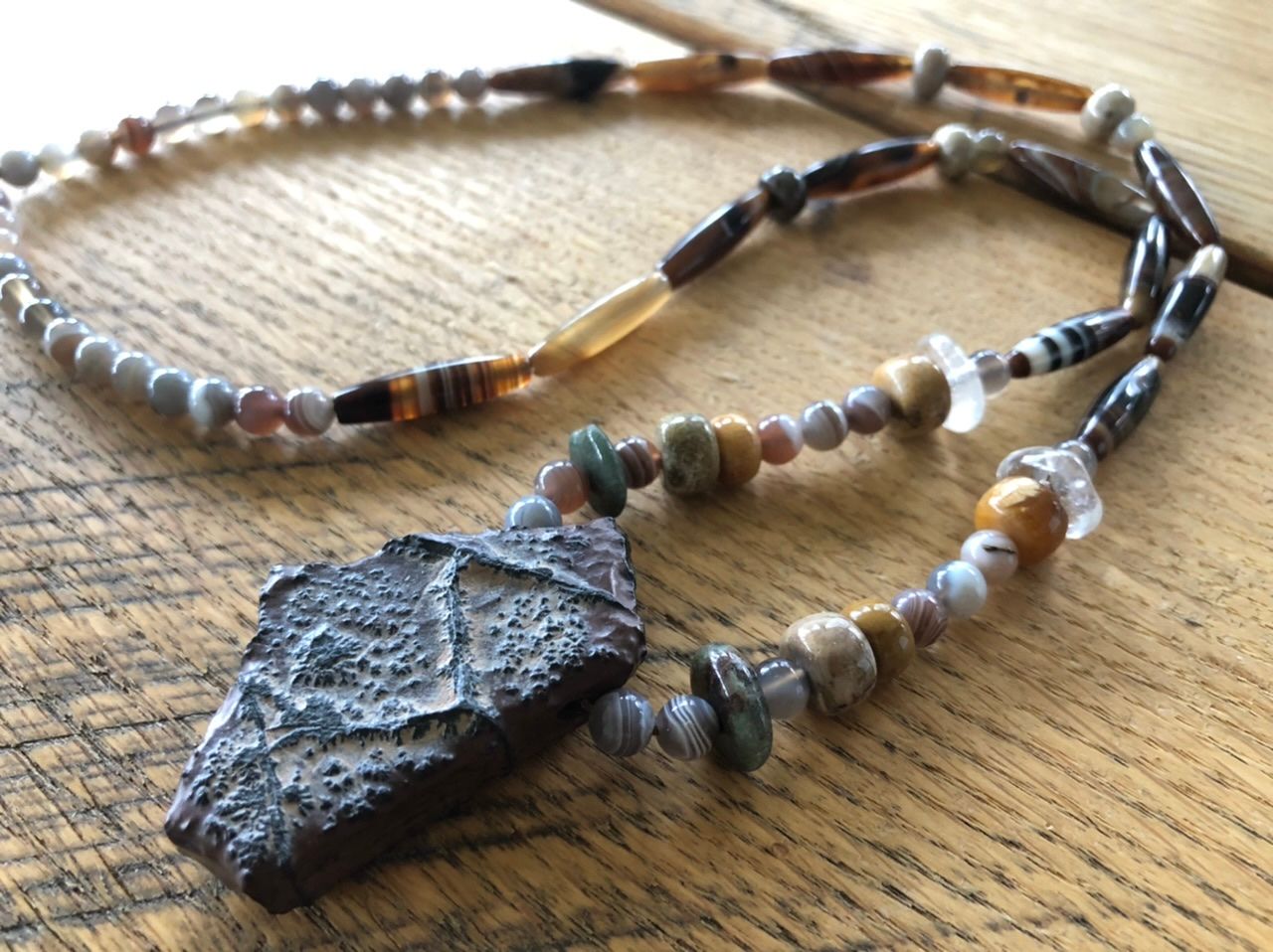 A necklace of earth tone agates and fossils rests on an oak table