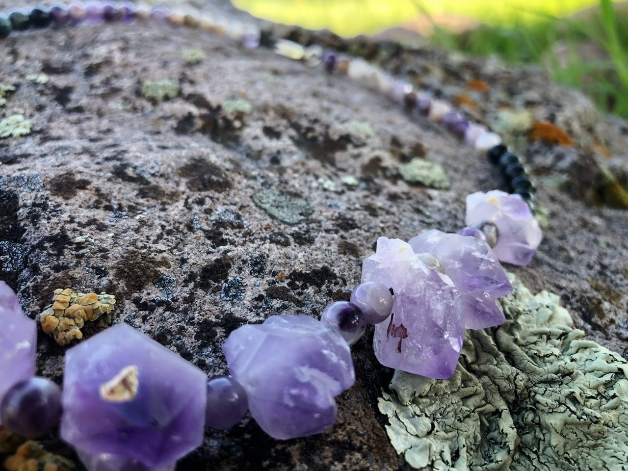 A purple amethyst crystal and black cats eye necklace rests on a lichen covered Boulder
