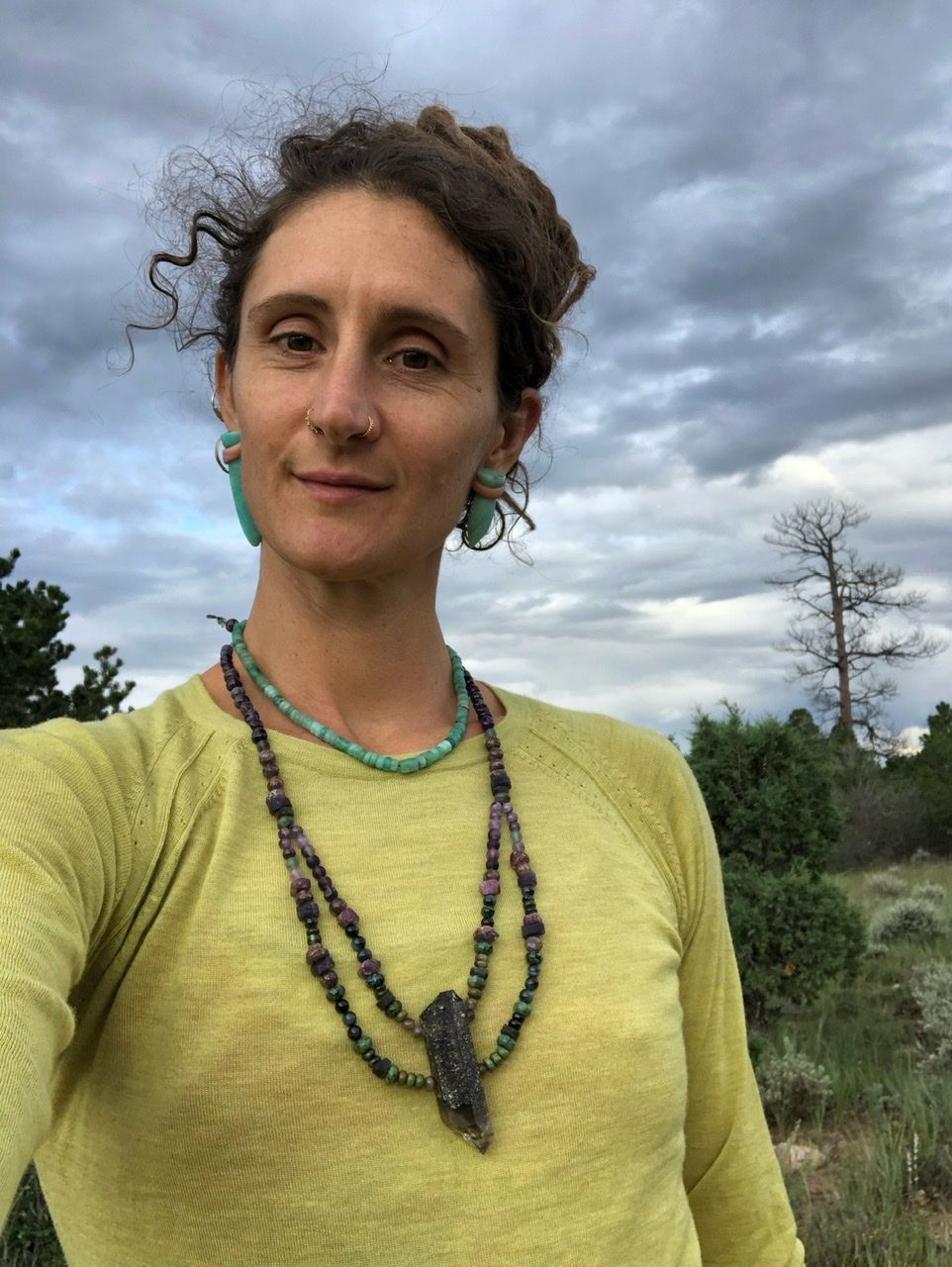 A woman in a chartreuse sweater, green stone earrings and necklace, wears a long crystal purple and green stone Necklace in the forest 