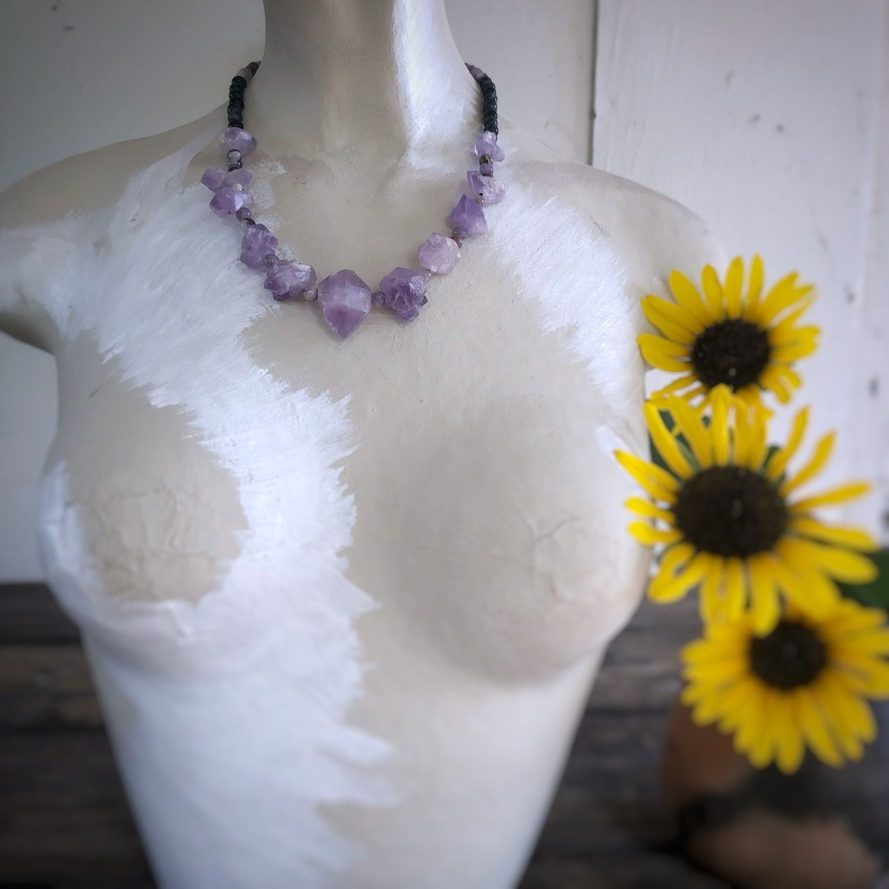 A amethyst crystal necklace rests on a white mannequin on a wood table with sunflowers in the background 