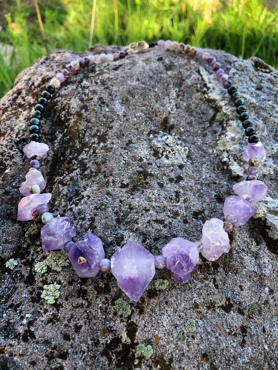 A purple amethyst crystal and black cats eye necklace rests on a lichen covered Boulder