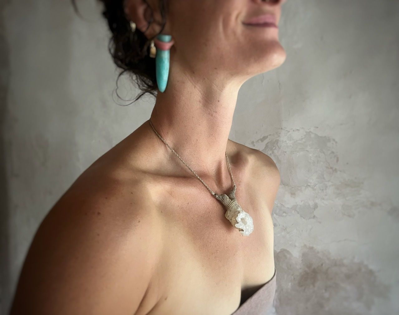 A woman wearing a pale purple top and a white thread and crystal necklace stands in front of a white mud wall