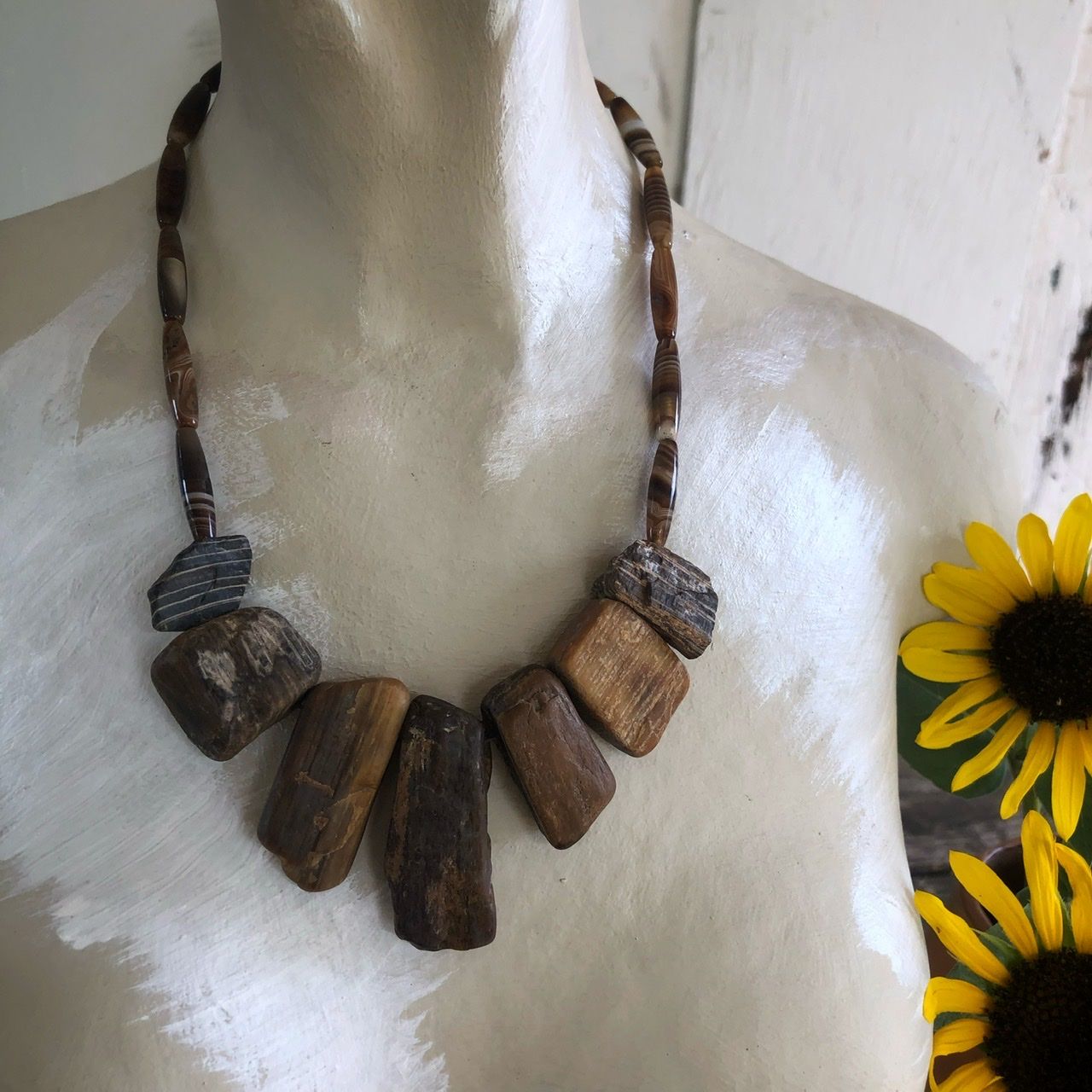 A petrified wood and banded agate necklace rests on a white mannequin won a wood table with sunflowers in the background as