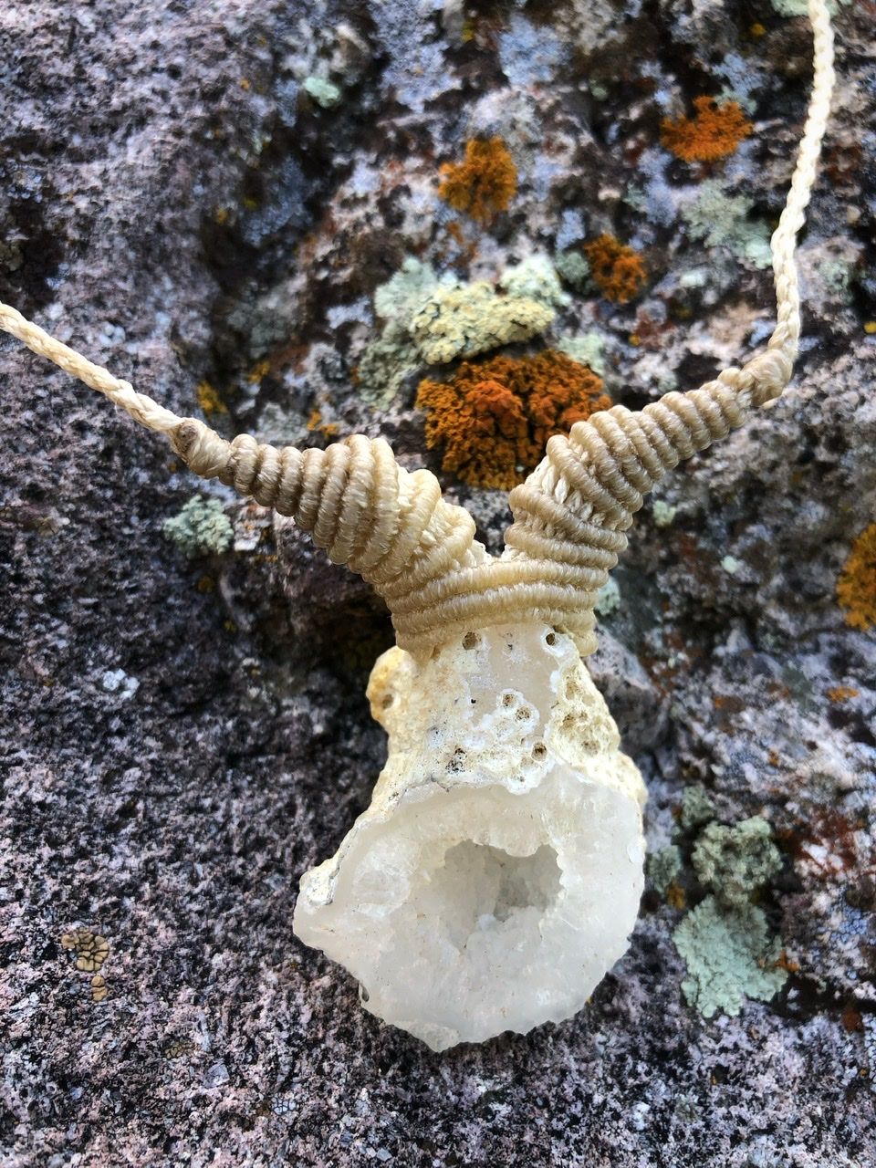 A small white geode and white knotwork string necklace rests on a lichen covered Boulder